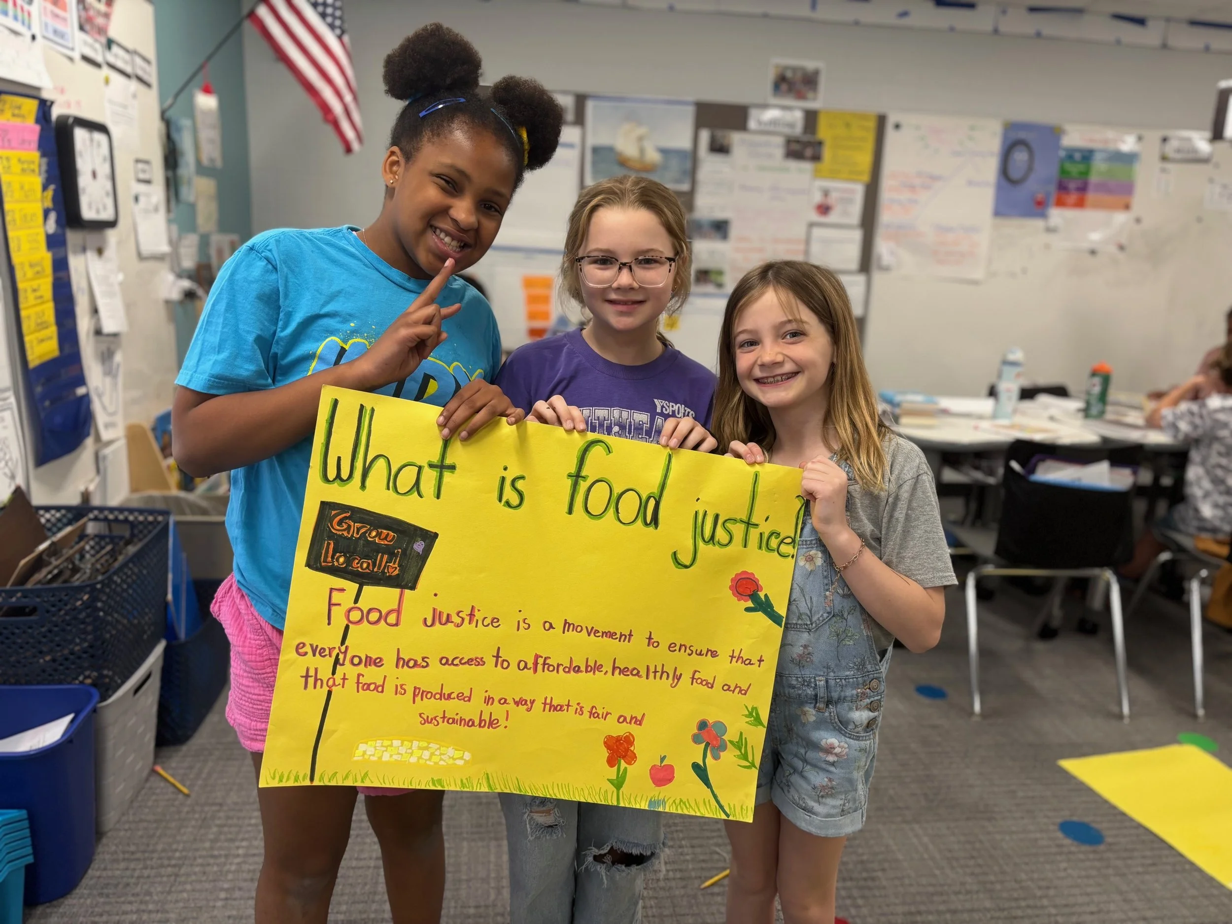 Three young girls in a classroom holding a yellow poster about food justice, with colorful drawings and handwritten text.