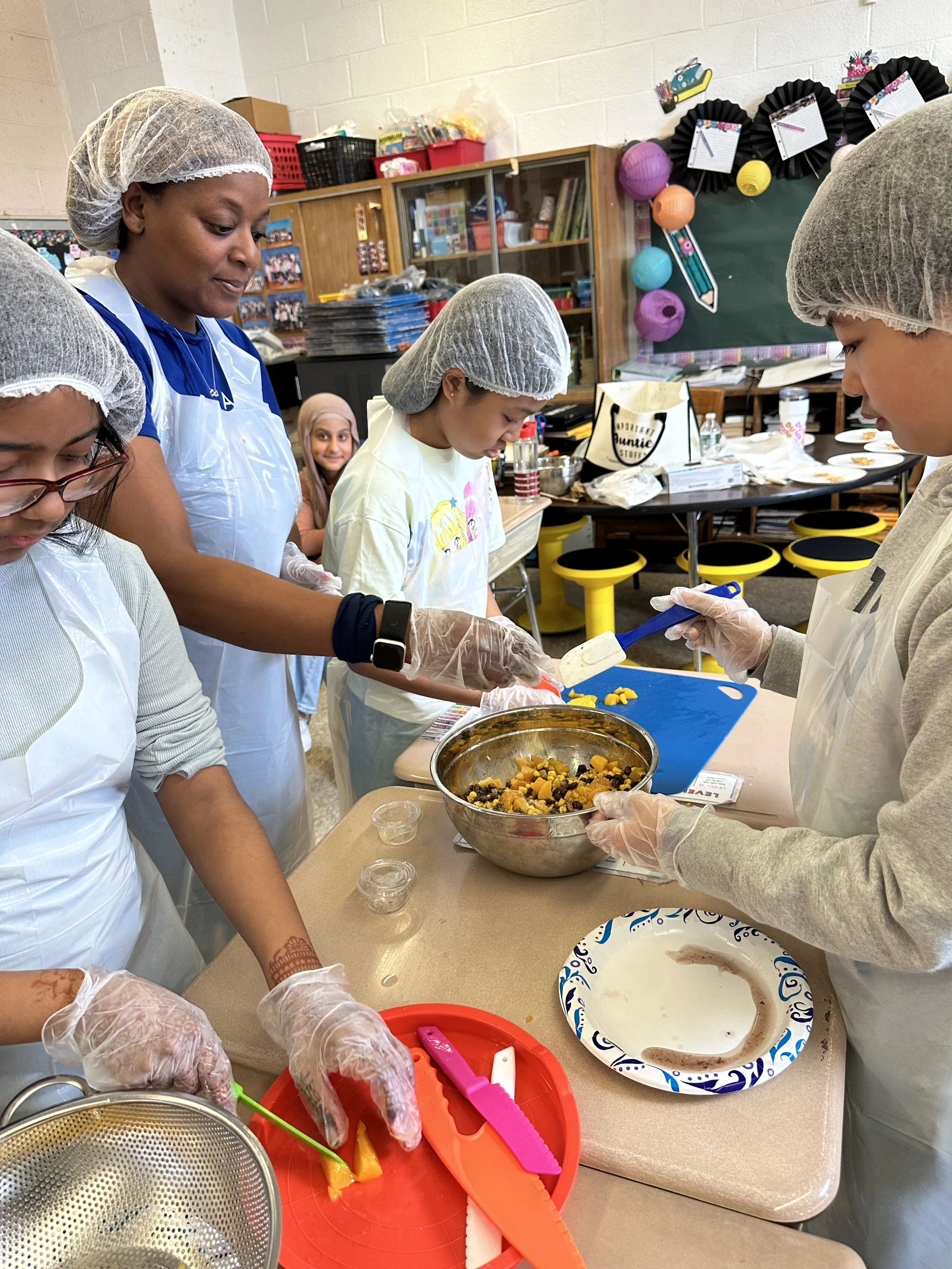 Group of children and an adult wearing hairnets and gloves, preparing food in a classroom. They are mixing ingredients in bowls and cutting vegetables, engaged in a cooking activity.