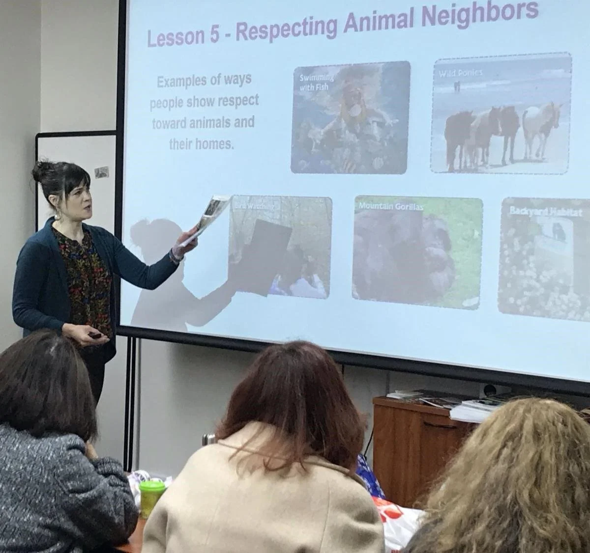 A man giving a presentation to a seated group of people in a conference room with large windows and a projection screen.