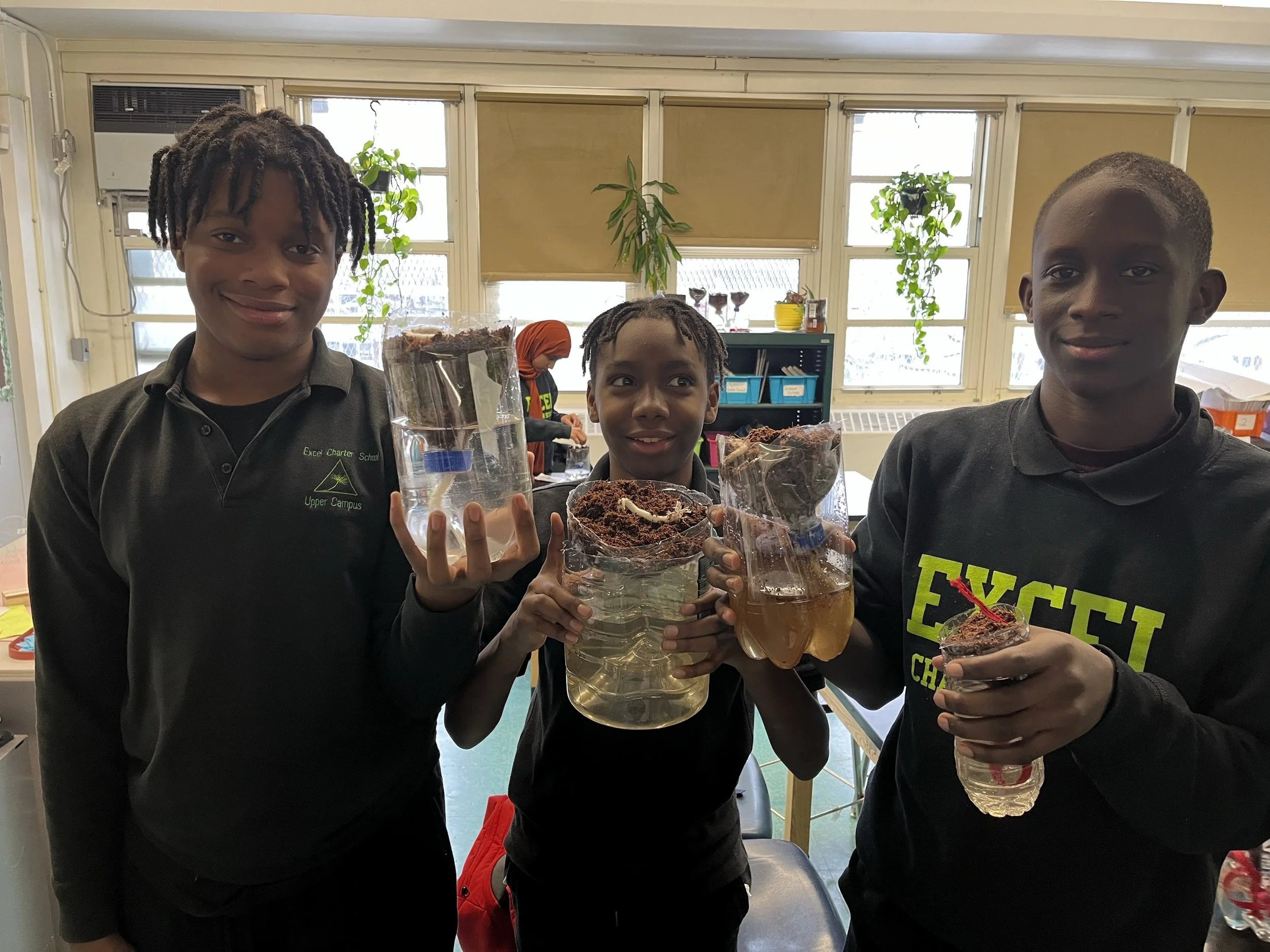 Three boys inside a classroom holding homemade volcano science projects made from plastic bottles, paper mache, and other materials, with one boy wearing a shirt that says 'Excel Charter School Upper Campus' and another wearing a shirt that says 'Evil C'.