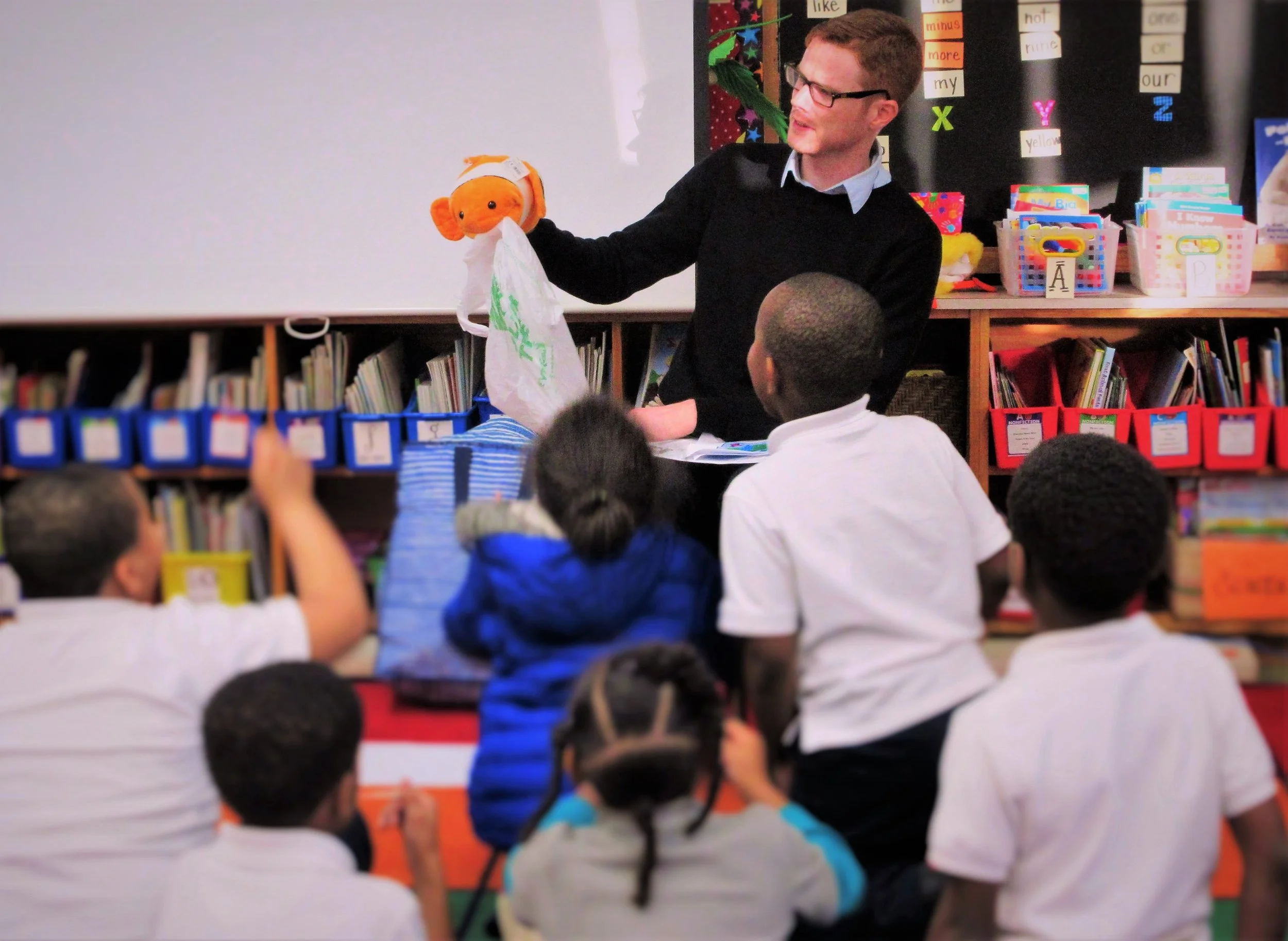 A teacher holding a puppet in front of a group of children in a classroom.