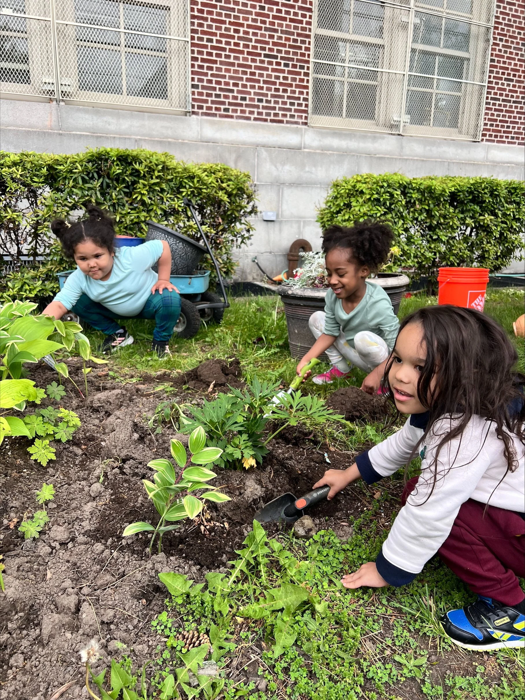 Three children planting and tending to plants in a garden in an urban area.
