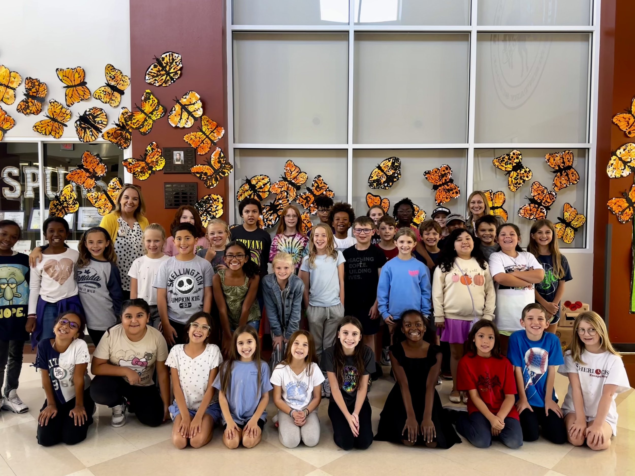 Group of children and two adults posing in front of butterfly decorations in a school or community center.