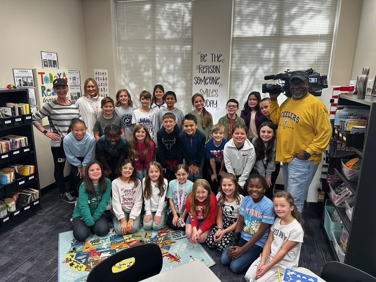 A group of children and adults in a library classroom, smiling for the camera. Two adults stand at each side, one with a video camera, while the children are in front, sitting and kneeling on a colorful rug with bookshelves and posters in the background.