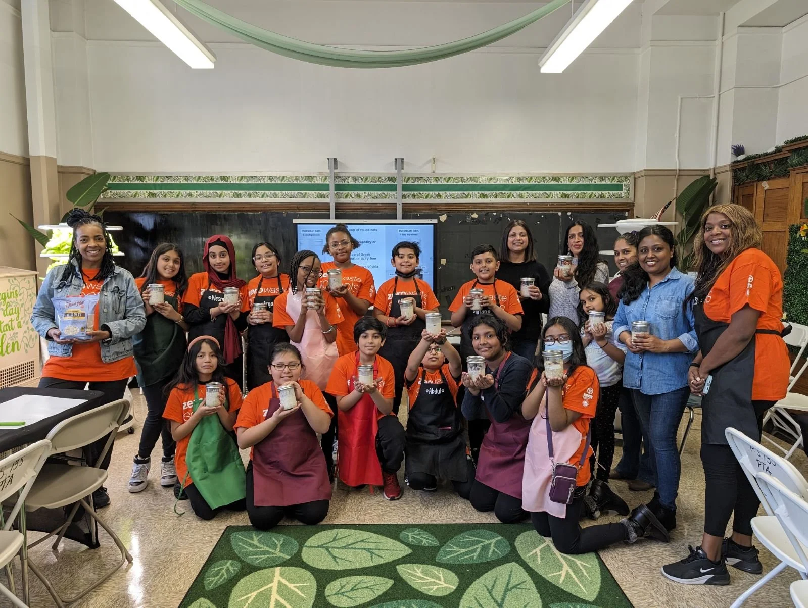 Group of children and adults in a classroom holding jars filled with oats, during a baking or cooking activity.