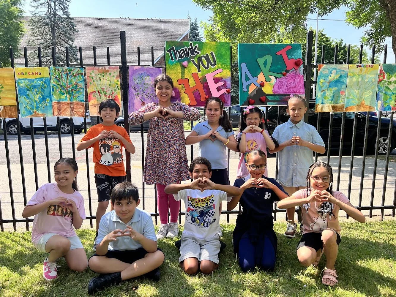 Group of children outdoors in front of fence displaying colorful artwork and a sign that says "Thank You The Art." They are posing with hands forming hearts, smiling, and standing on grass on a sunny day.