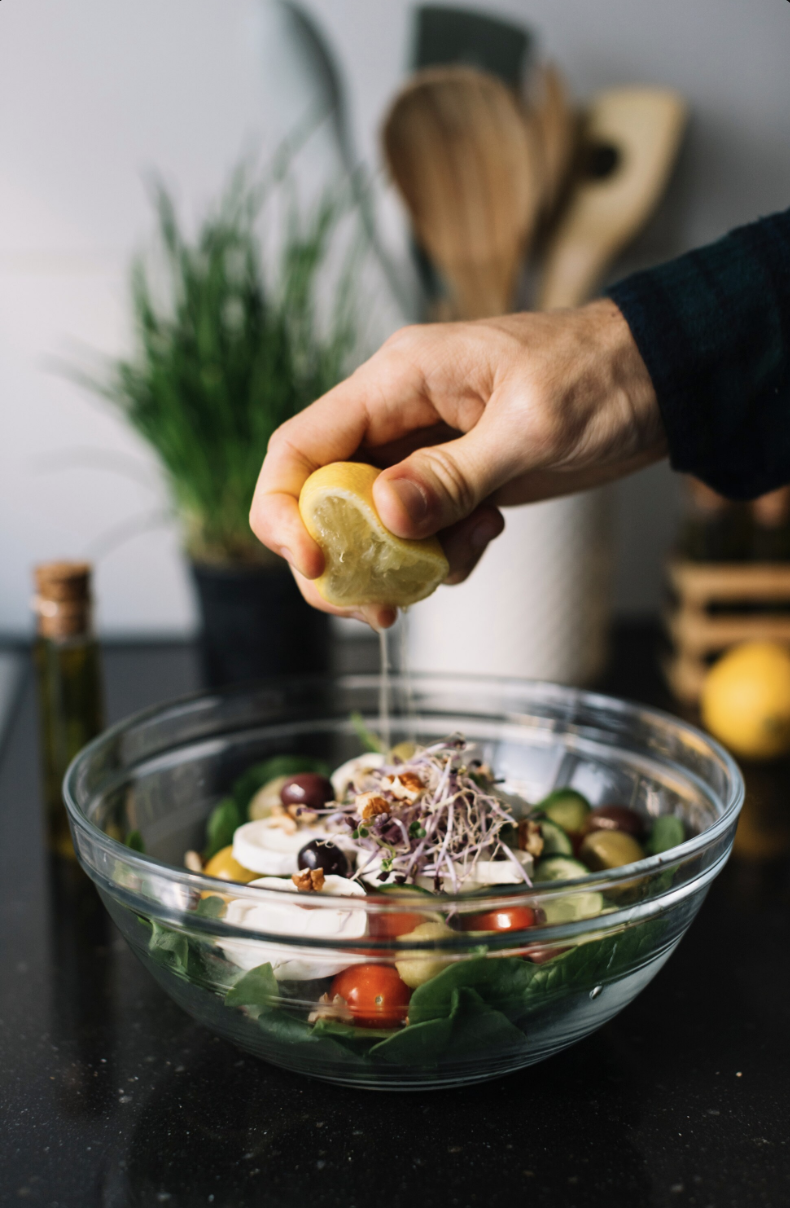 A person squeezing lemon over a fresh vegetable salad in a glass bowl on a black countertop, with kitchen utensils and a plant in the background.