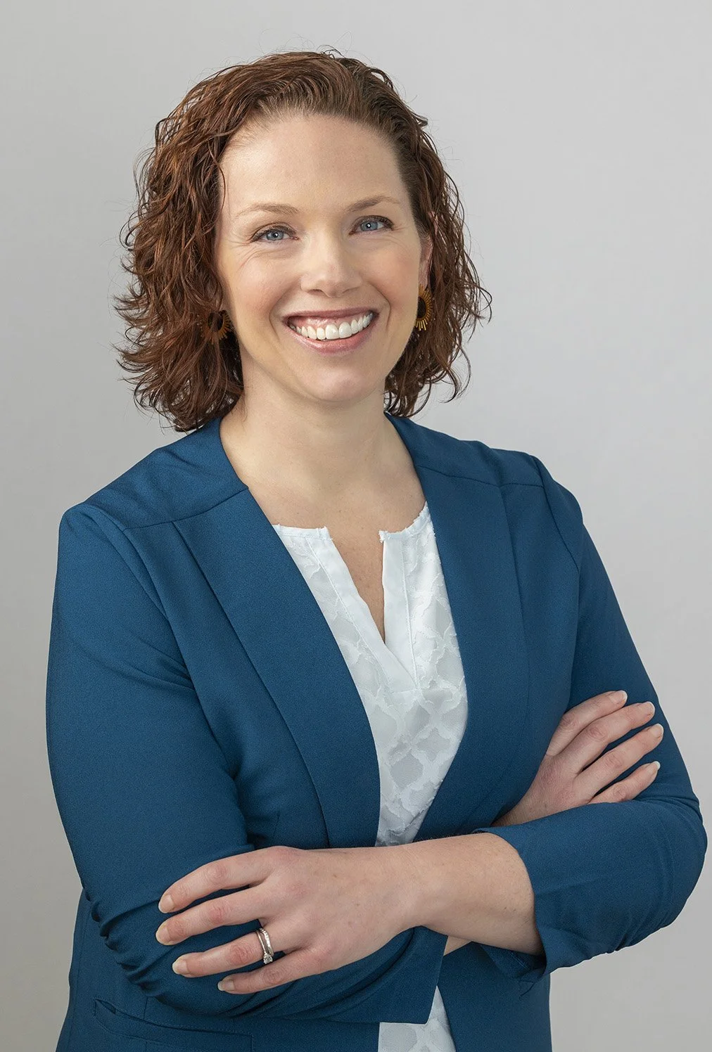 A woman with curly red hair wearing a teal blazer and white blouse, smiling with arms crossed against a plain light gray background.