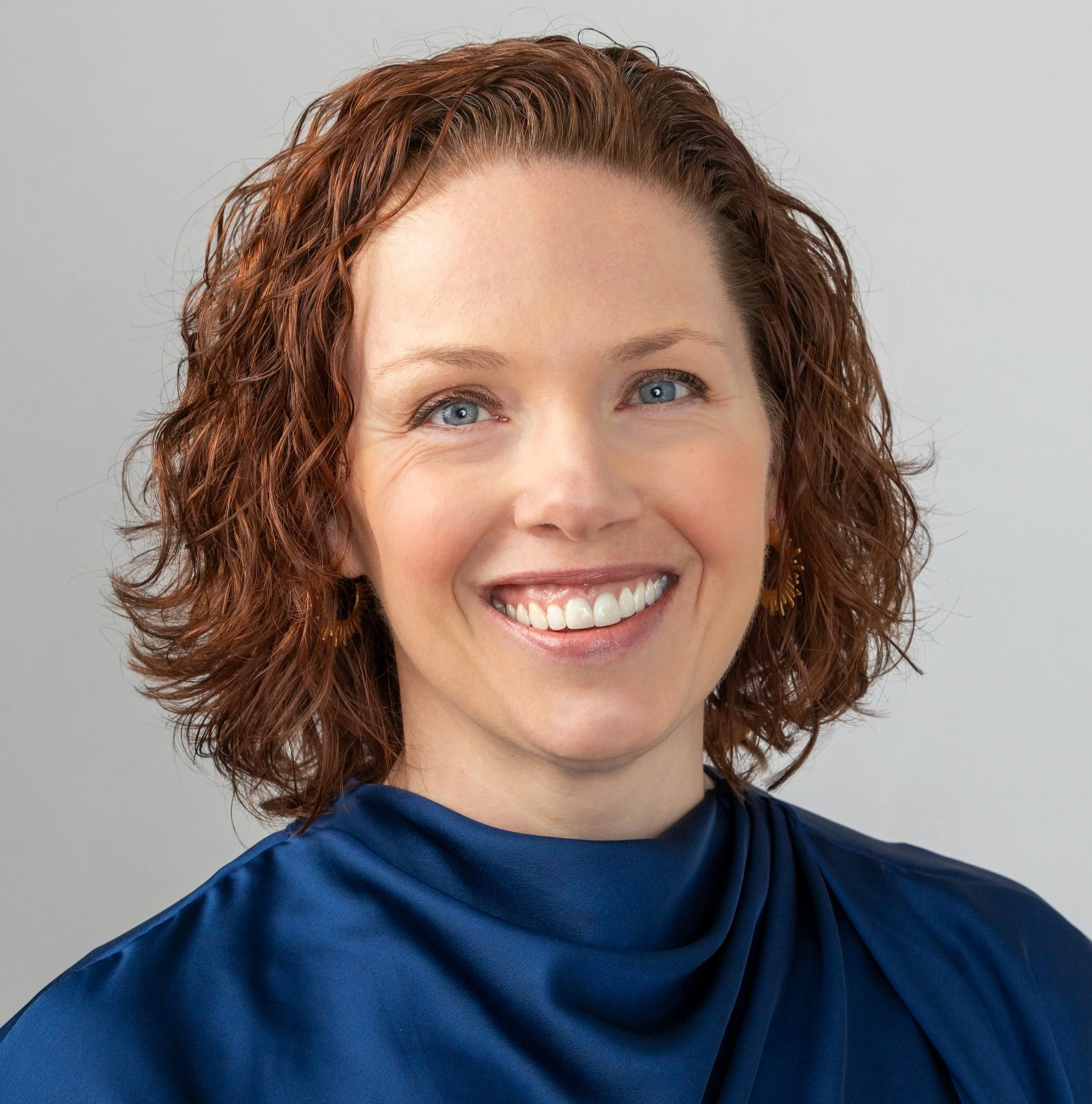 A woman with curly red hair, blue eyes, and a broad smile. She is wearing a blue top and gold earrings, with a plain gray background.