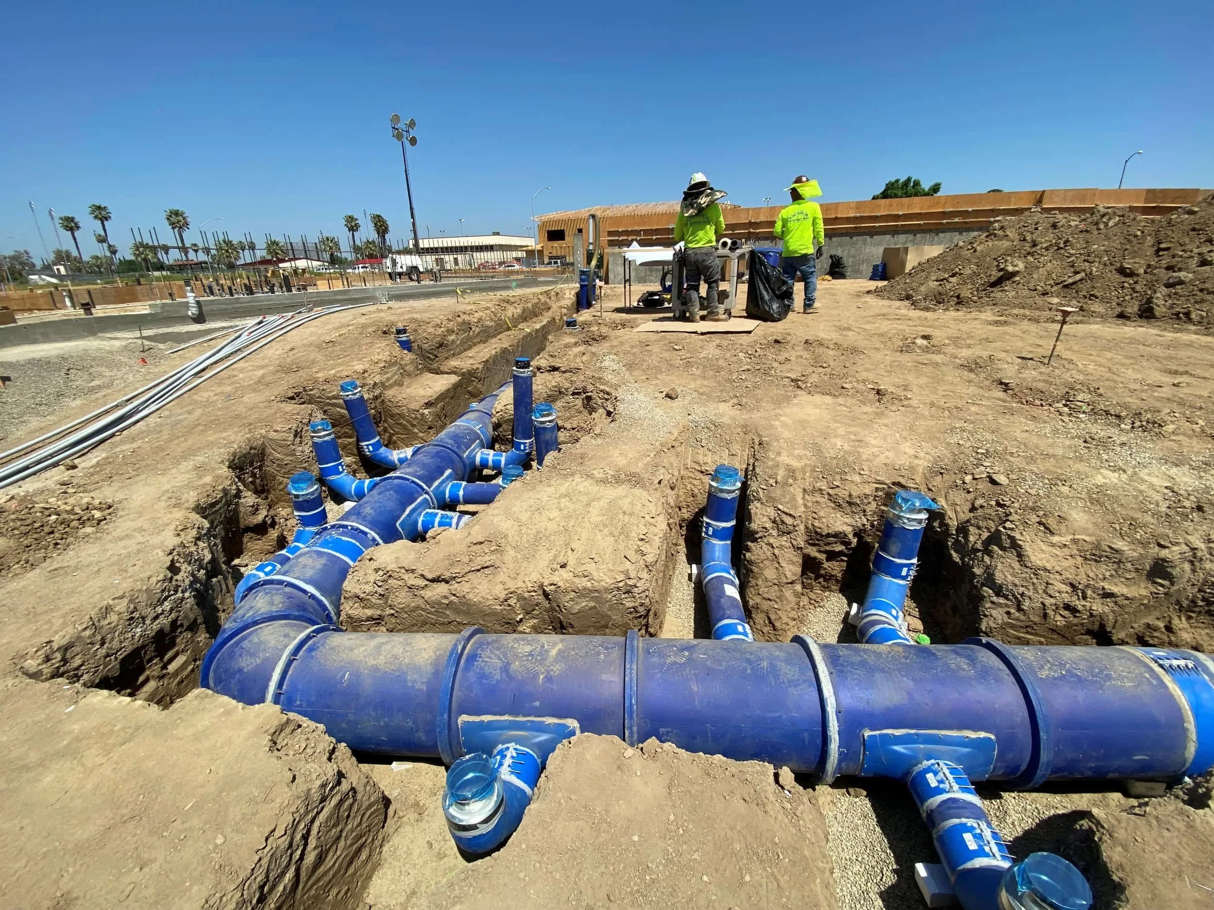 Construction workers in yellow safety vests and hats working on underground HVAC pipes on a construction site under a clear blue sky.