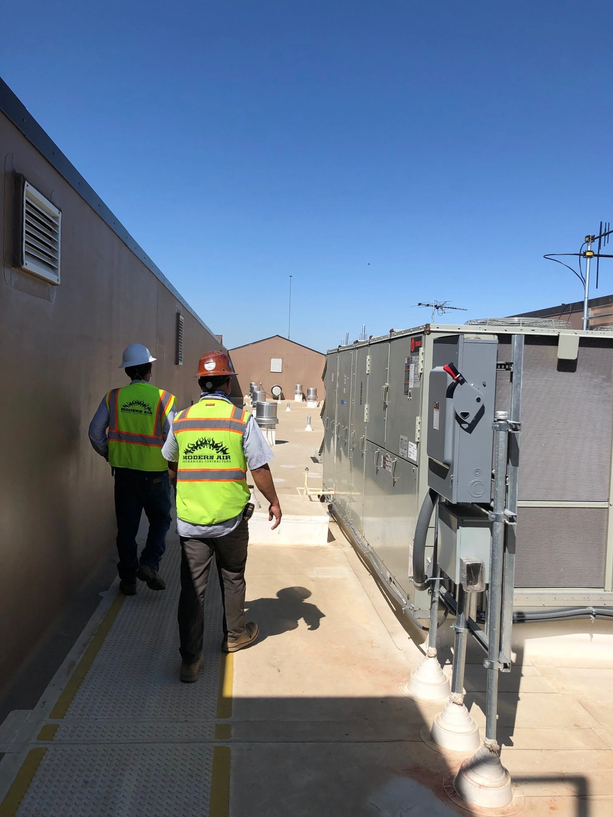 Two construction workers wearing safety vests and helmets walking on a rooftop with HVAC units and vents.