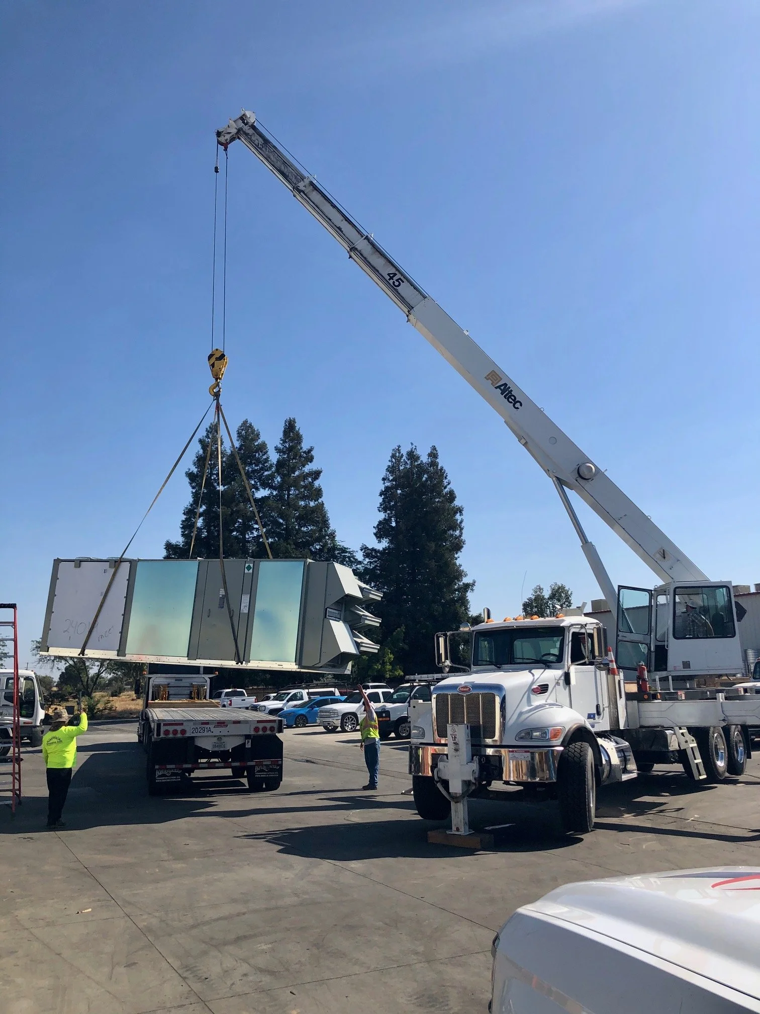 A crane lifting a large metallic HVAC unit in a parking lot with several workers and parked cars, some trees in the background.