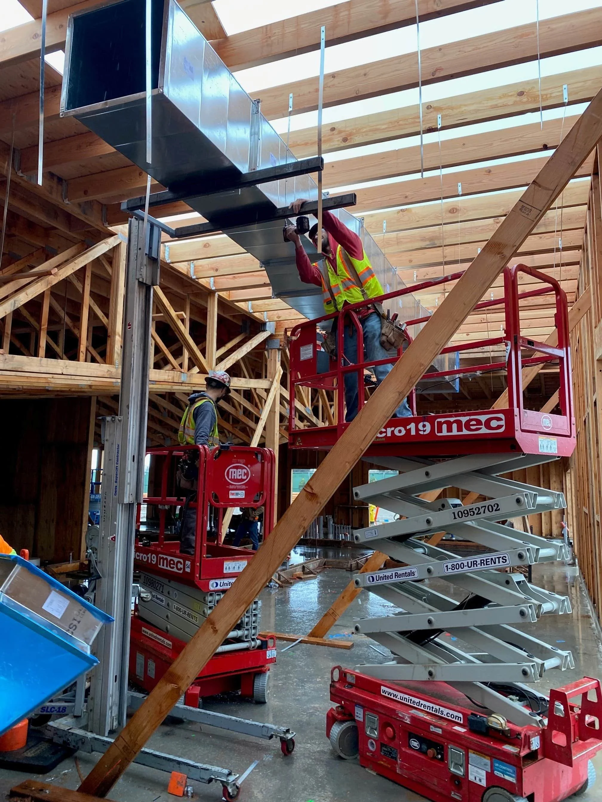 Construction workers installing ductwork on the ceiling of a building under construction, using a scissor lift and wearing safety gear.