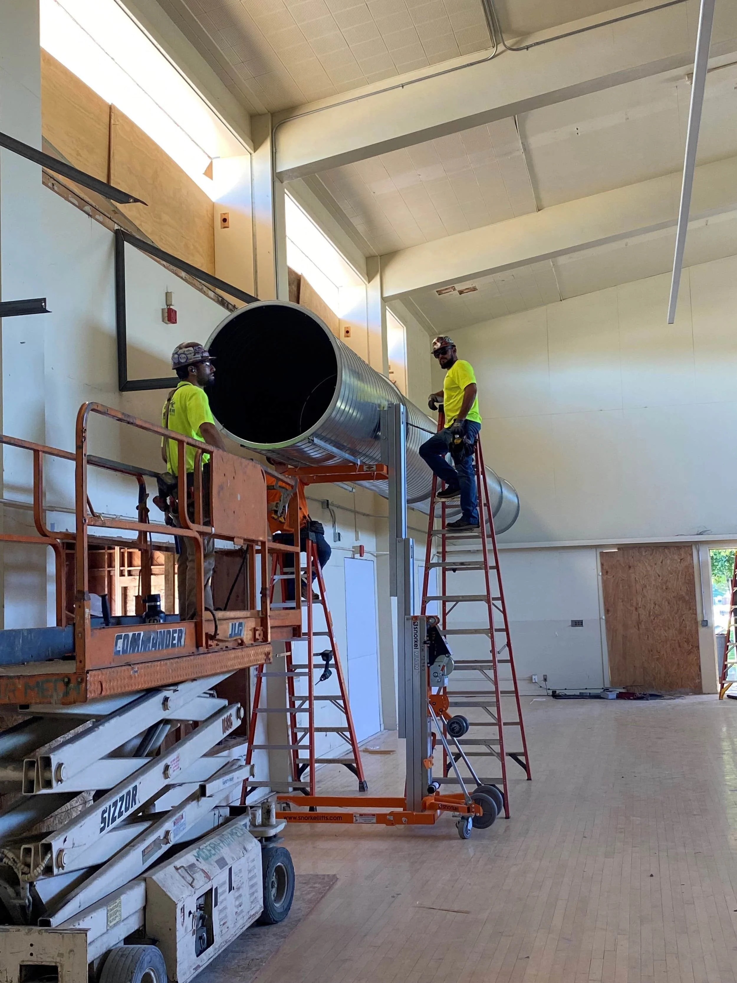 Two construction workers installing or repairing ductwork inside a building. One is standing on a lift, and the other is on a ladder, both wearing yellow safety shirts and helmets.
