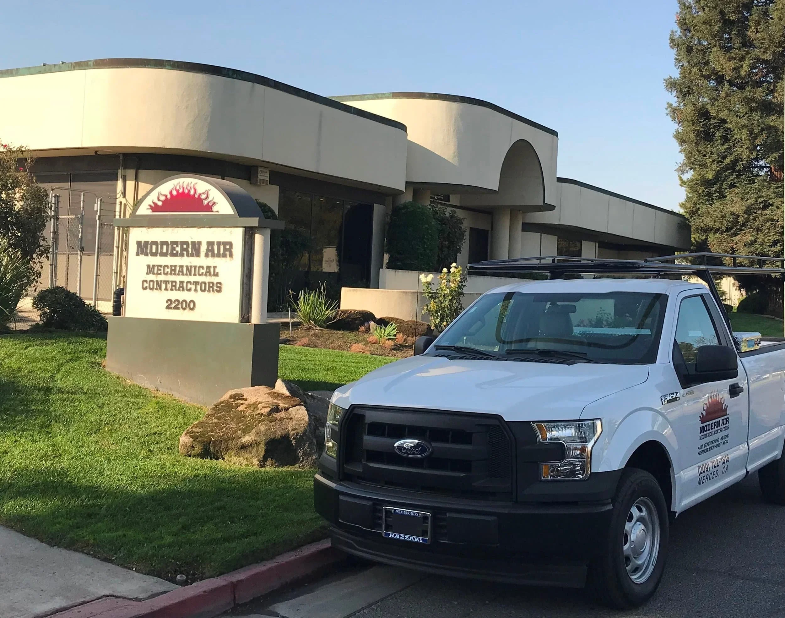 Front view of a commercial building with a sign reading 'Modern Air Mechanical Contractors, 2200'. A white Ford work truck with the same company's logo and contact information is parked in front. The building has a curved architectural design with bushes and rocks in the landscaping.