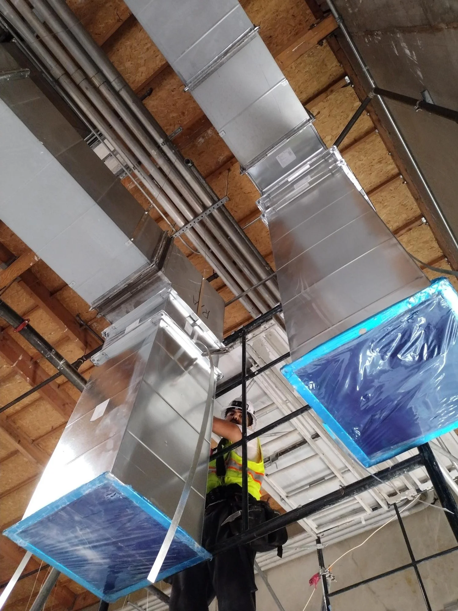 Construction worker in safety vest and helmet installing or repairing metal HVAC ductwork on ceiling of a building.