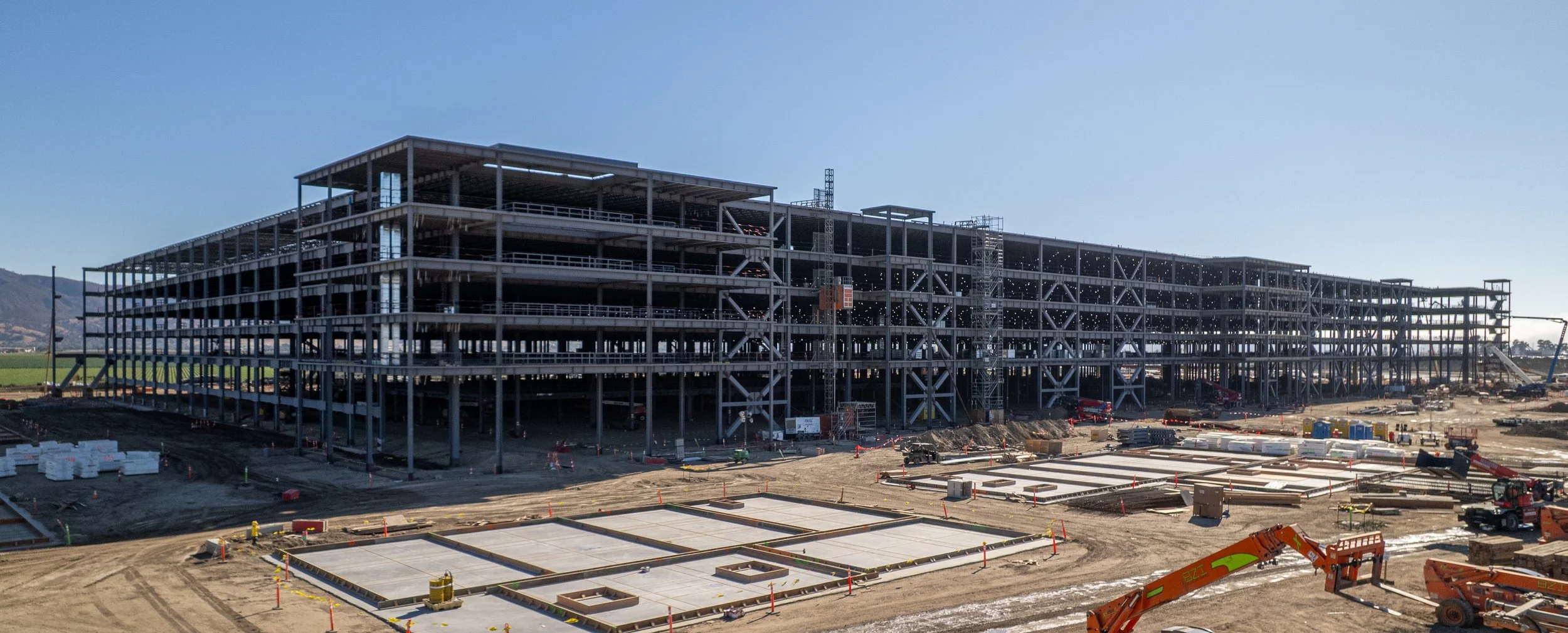 Construction site with a large multi-story steel frame building under construction, several construction vehicles and materials on the ground, and mountains in the background.