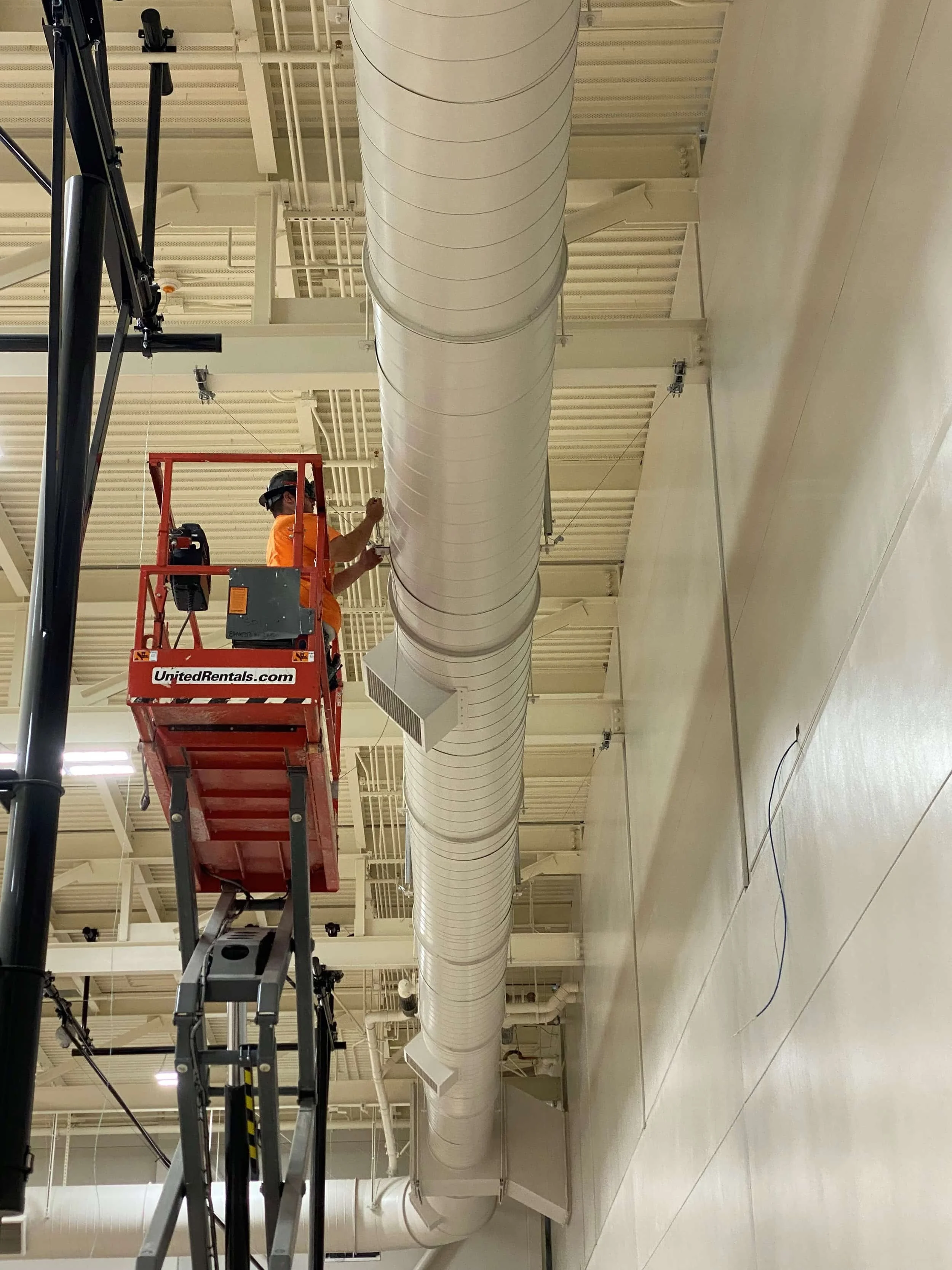 A worker standing on a red lift platform cleaning or inspecting a large white industrial air duct in a commercial or industrial building.