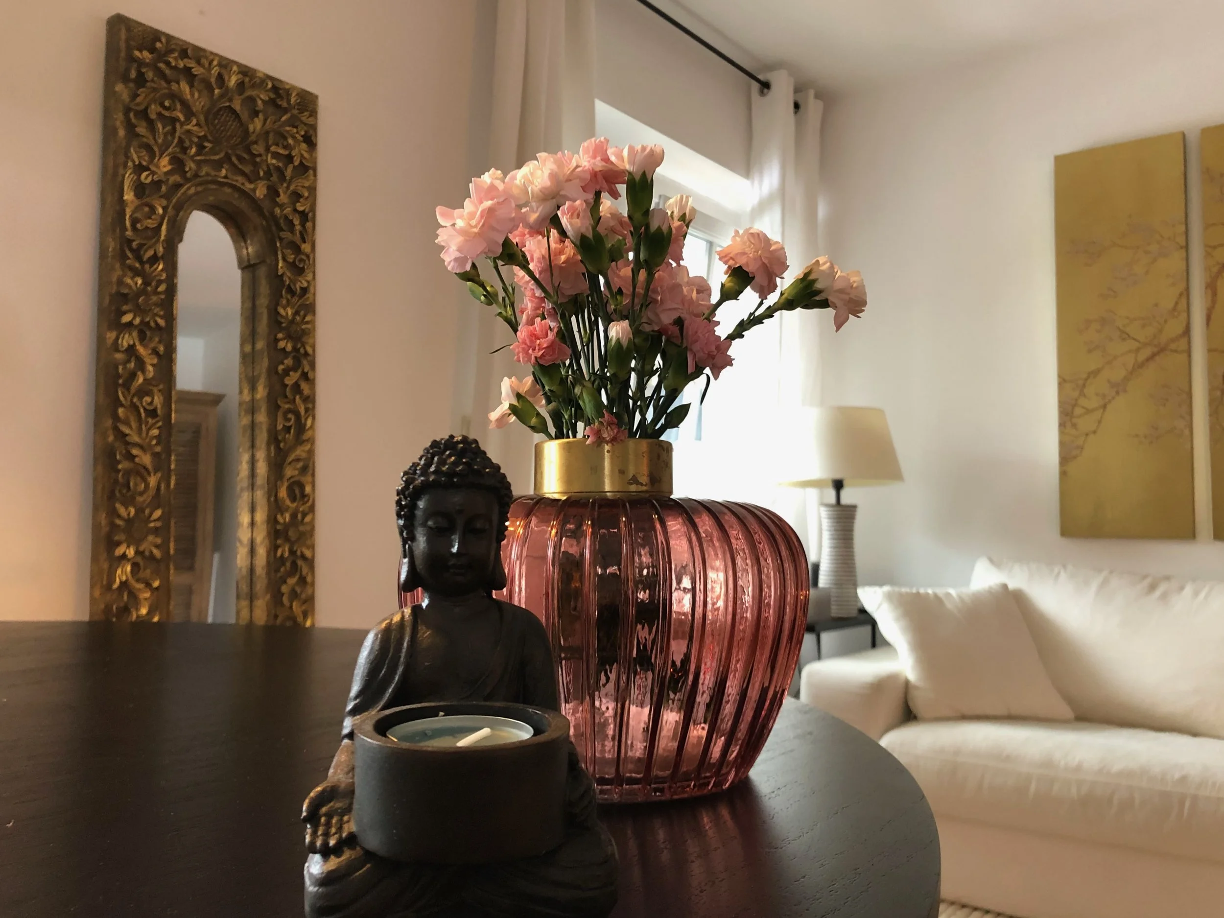 Vase mit rosa Blumen, eine Buddha-Statue, eine rote Mid-Century Vasen, Kerze, Wandkunst und Stehlampe in einer modernen Wohnzimmergestaltung.