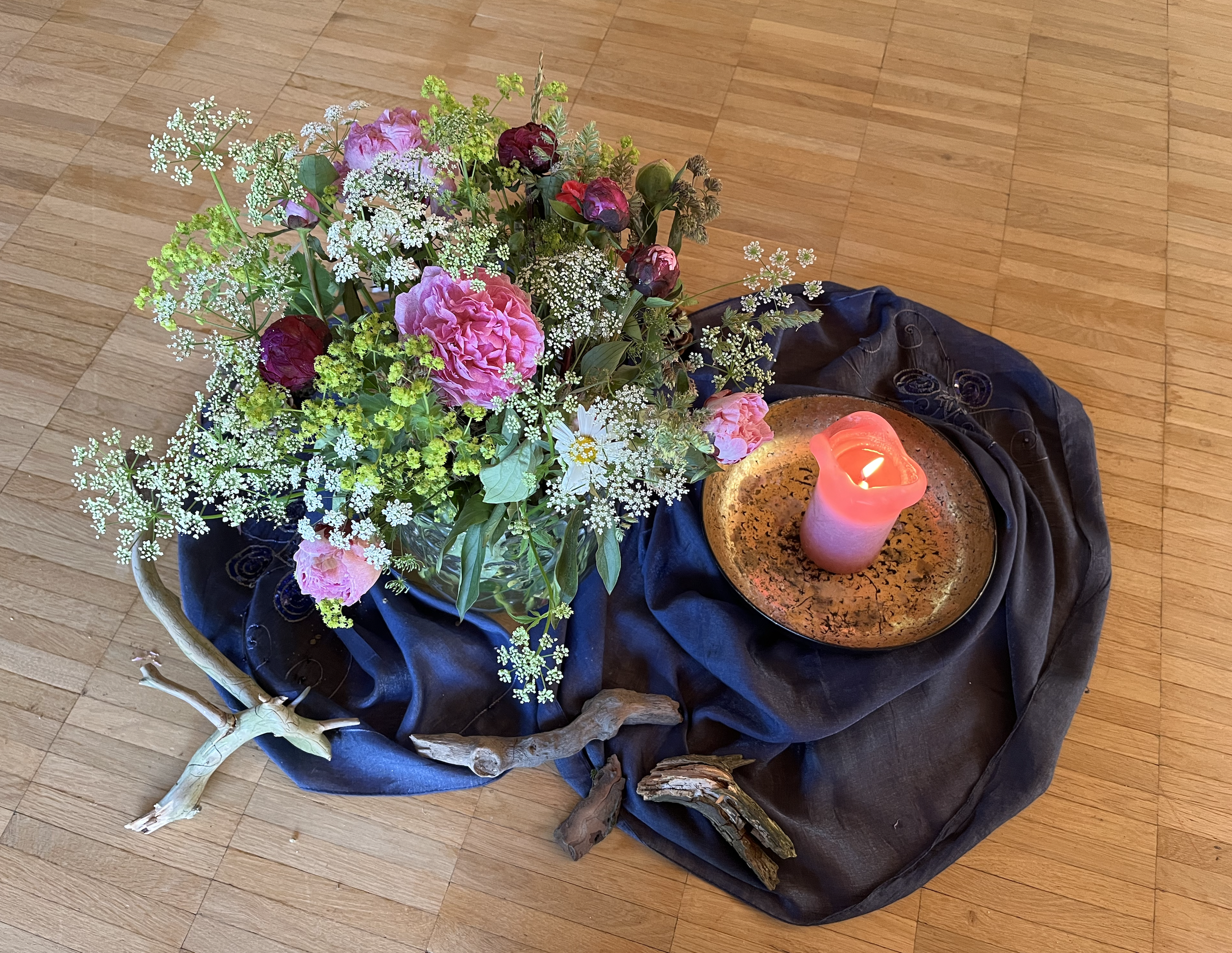 blumenstrauß mit pinken und roten blumen, weißen kleinen blumen und grünem blattwerk auf einem schwarzen tuch, daneben eine brennende rosa kerze auf einer steinplatte, auf holzparkett