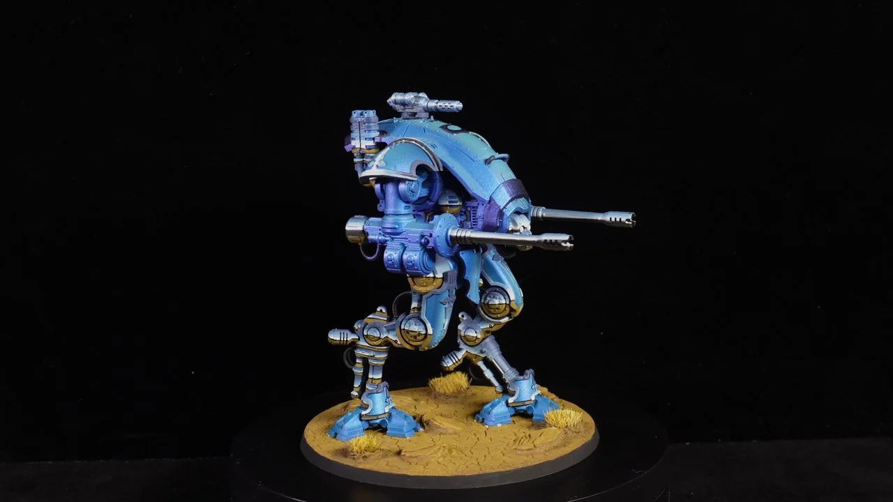 A detailed miniature model of a futuristic, blue mech robot with two long guns, standing on a desert terrain base with dry grass patches, against a black background.