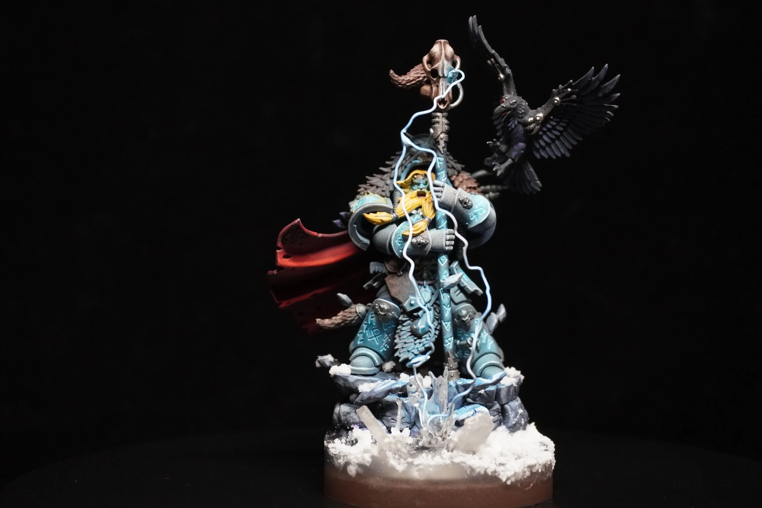 A detailed miniature figurine depicts a fantasy warrior in ornate armor, holding a staff, with a bird perched on his shoulder and a skull atop his helmet, against a black background.