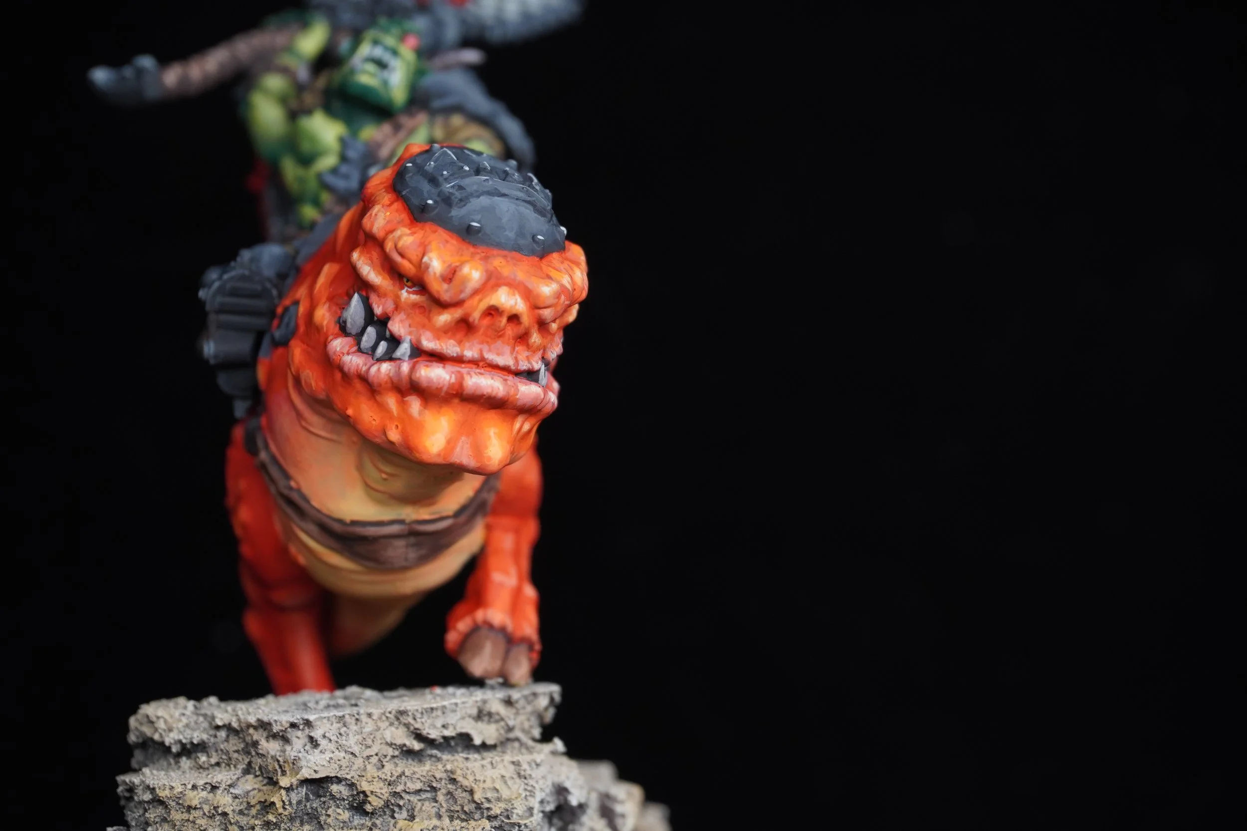 A detailed figurine of a monstrous creature with an orange face, black and gray head, sharp teeth, and a large belly, standing on a rough textured surface against a black background.