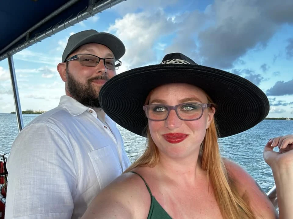 A man and woman taking a selfie on a boat with water and a partly cloudy sky in the background. The woman is wearing a large black hat, red lipstick, and glasses, and the man is wearing a gray cap and glasses.