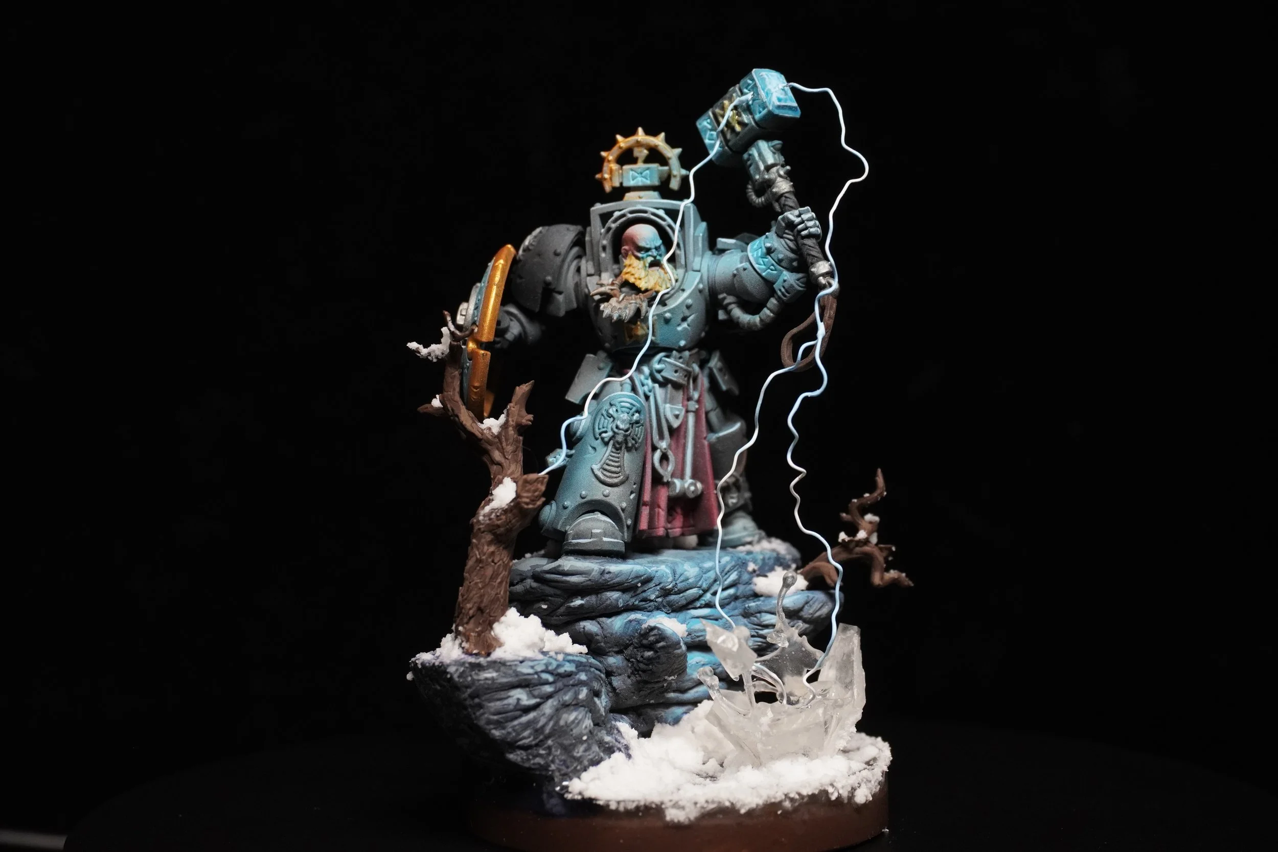 A detailed miniature figure of a sci-fi warrior in armor, standing on a rocky snowy base with twisted trees and ice formations, depicted with lightning effects.