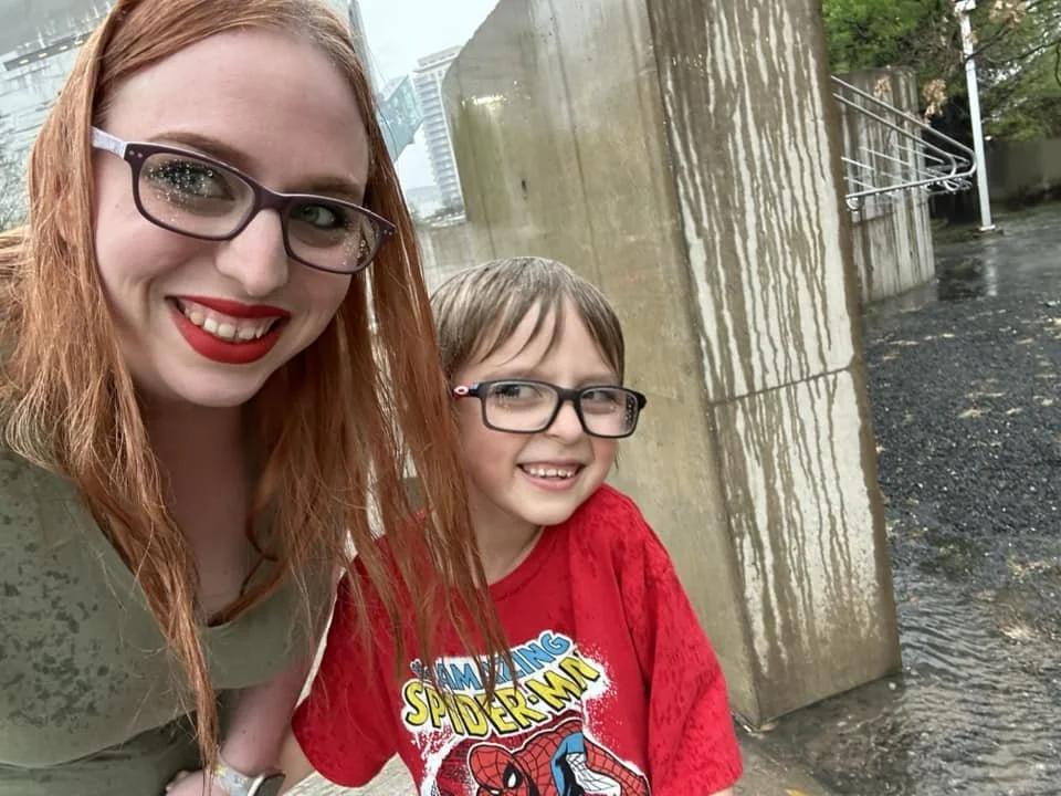 A woman and a young boy smiling outdoors in front of a concrete water structure, with the woman wearing glasses, red lipstick, and a beige shirt, and the boy wearing glasses and a red Spider-Man shirt.