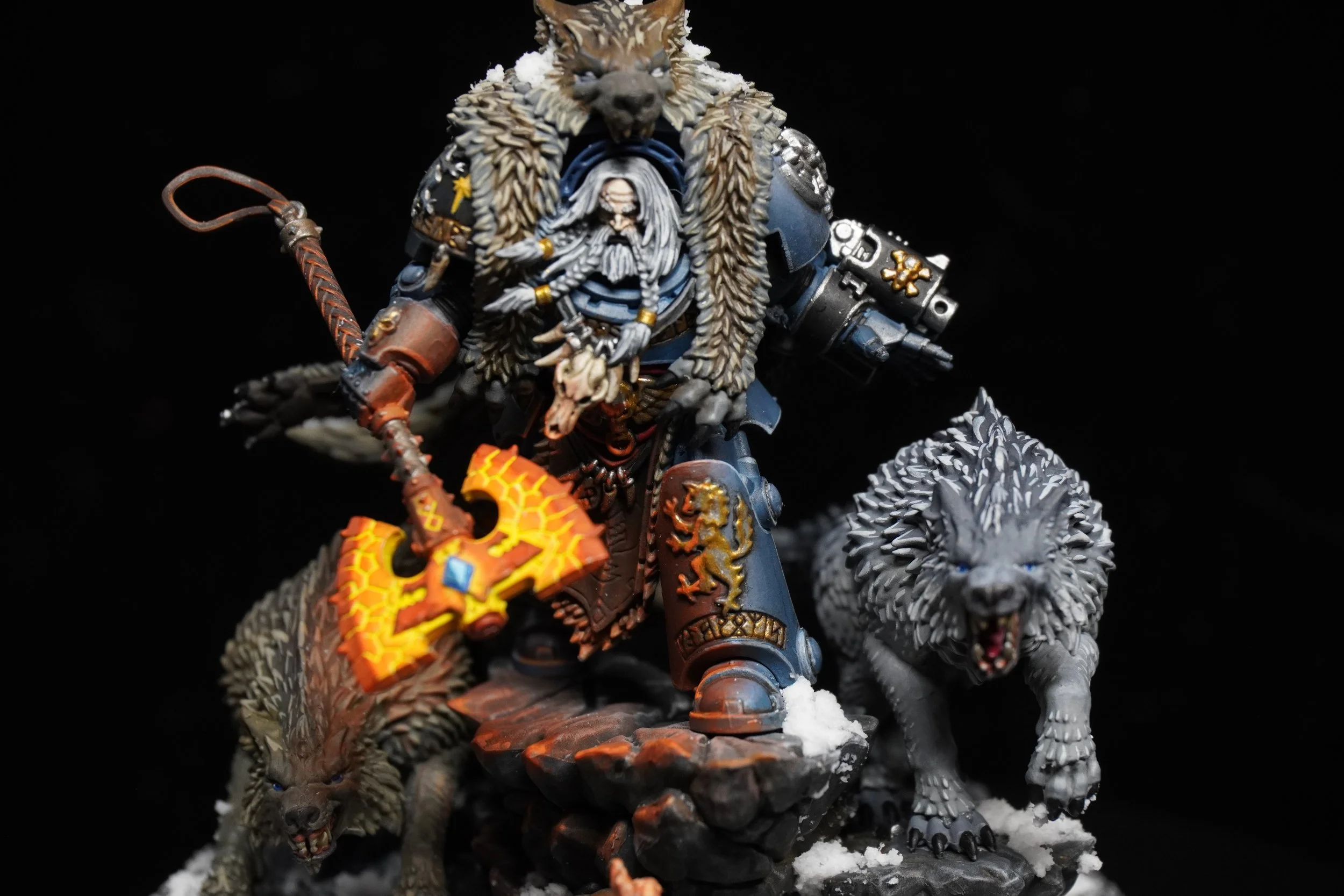 A highly detailed miniature figurine of a warrior with a wolf head helmet, wearing armor decorated with lion emblems, holding a fiery axe, and accompanied by two snarling wolves on a snowy base.
