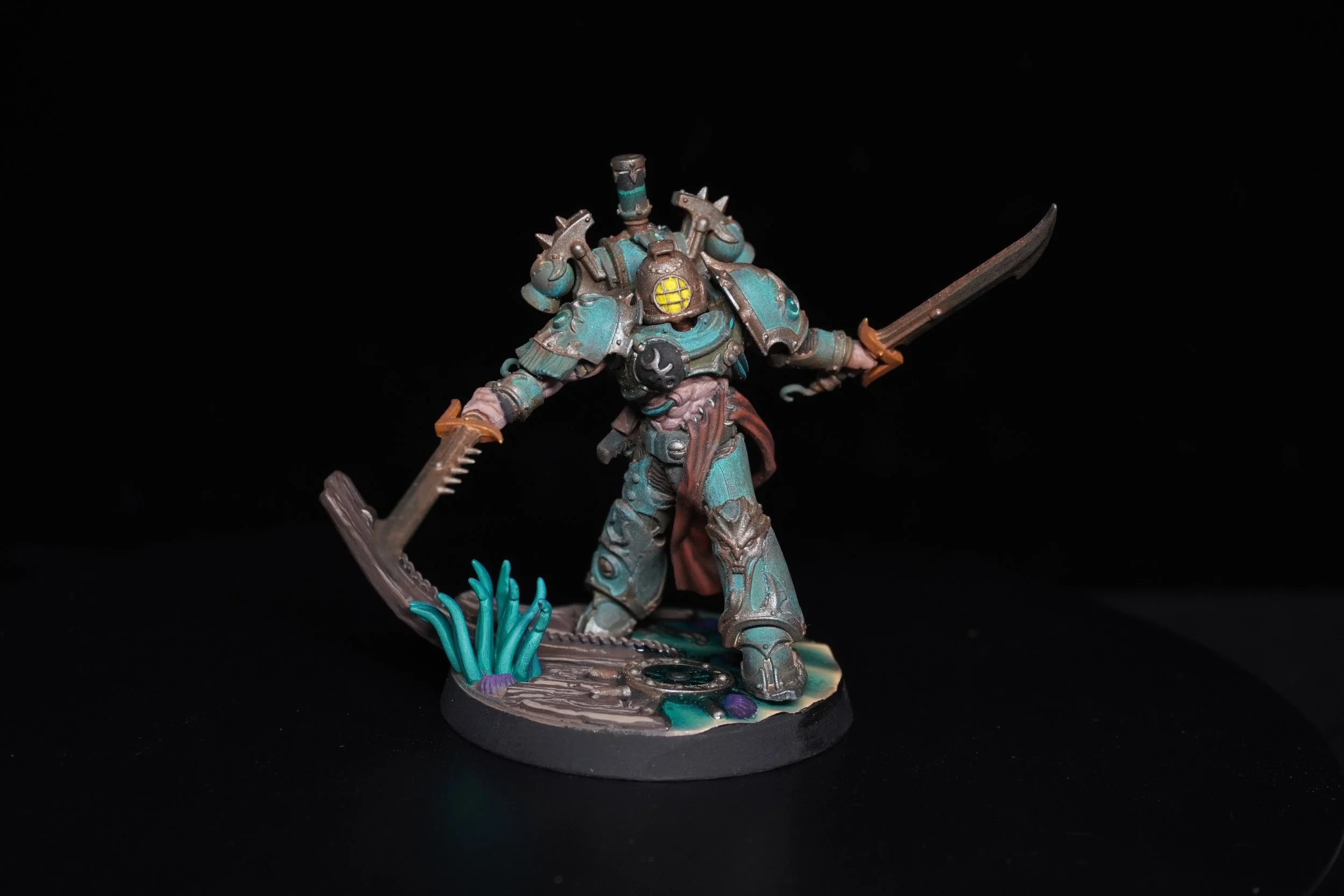 Miniature warrior figurine with armor, holding a sword and a saw, standing on a detailed base with plants and water elements, against a black background.