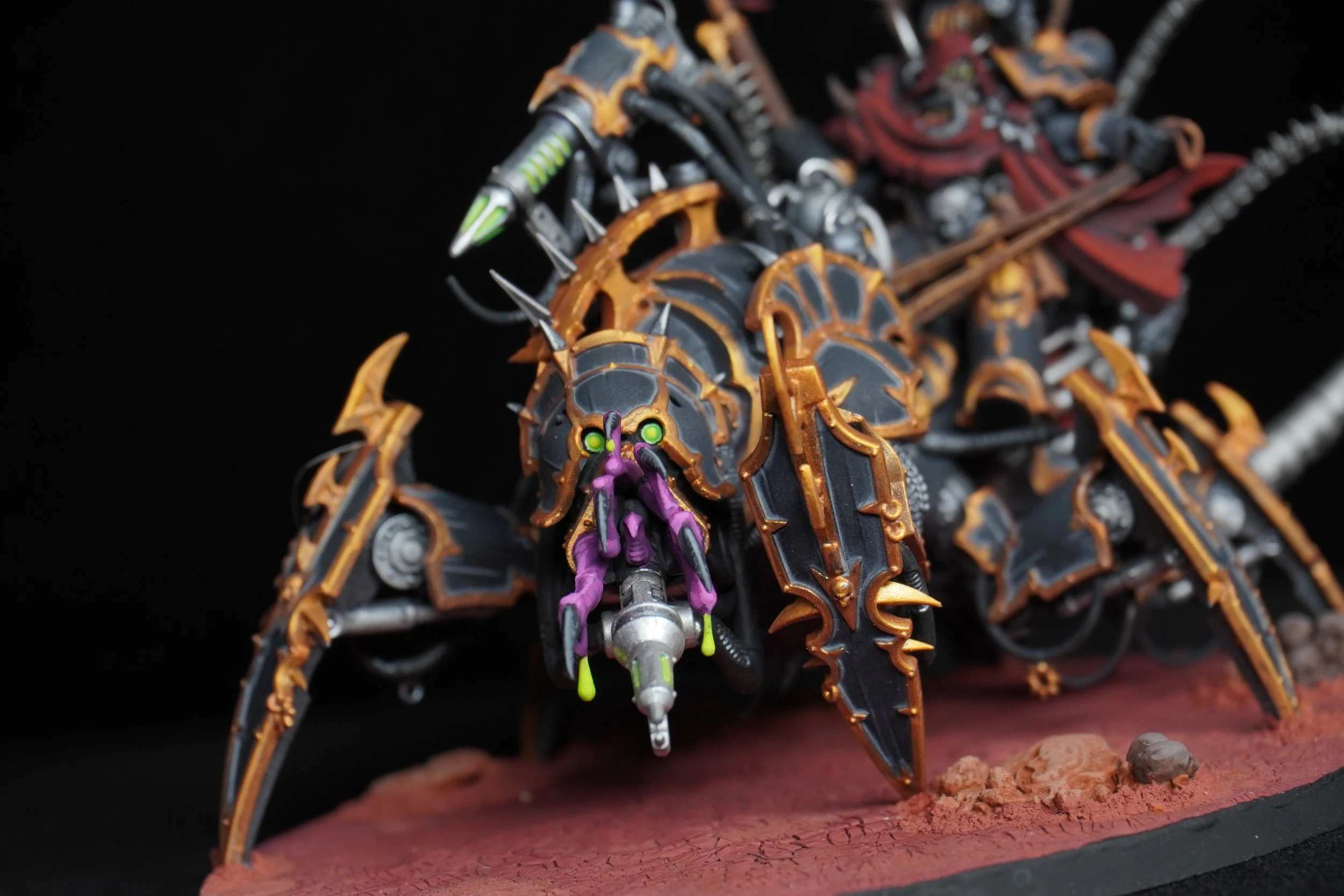 A detailed custom painted miniature model of a spider-like creature with mechanical and sci-fi elements, black and gold armor, green and purple accents, and a weapon mounted on its front, set on a textured red base.