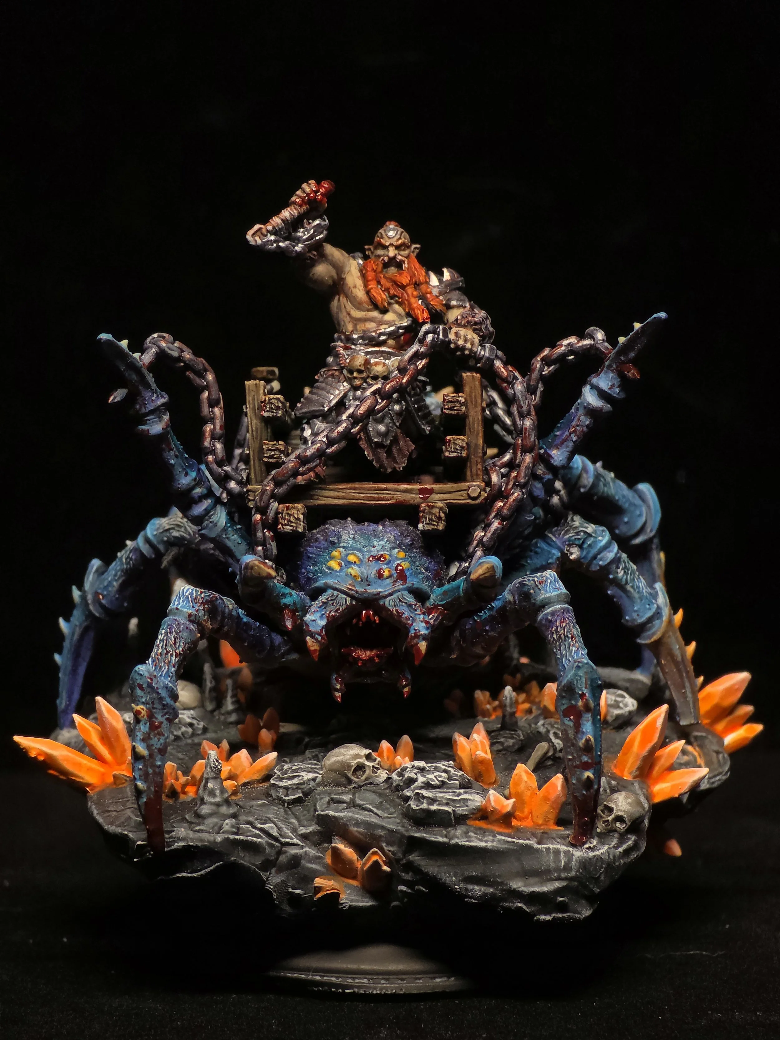 A detailed painted miniature of a fantasy scene featuring a large, multi-eyed spider with an armored humanoid figure riding on top, wielding a weapon. The scene is set on a rocky base with orange crystals and skull decorations.