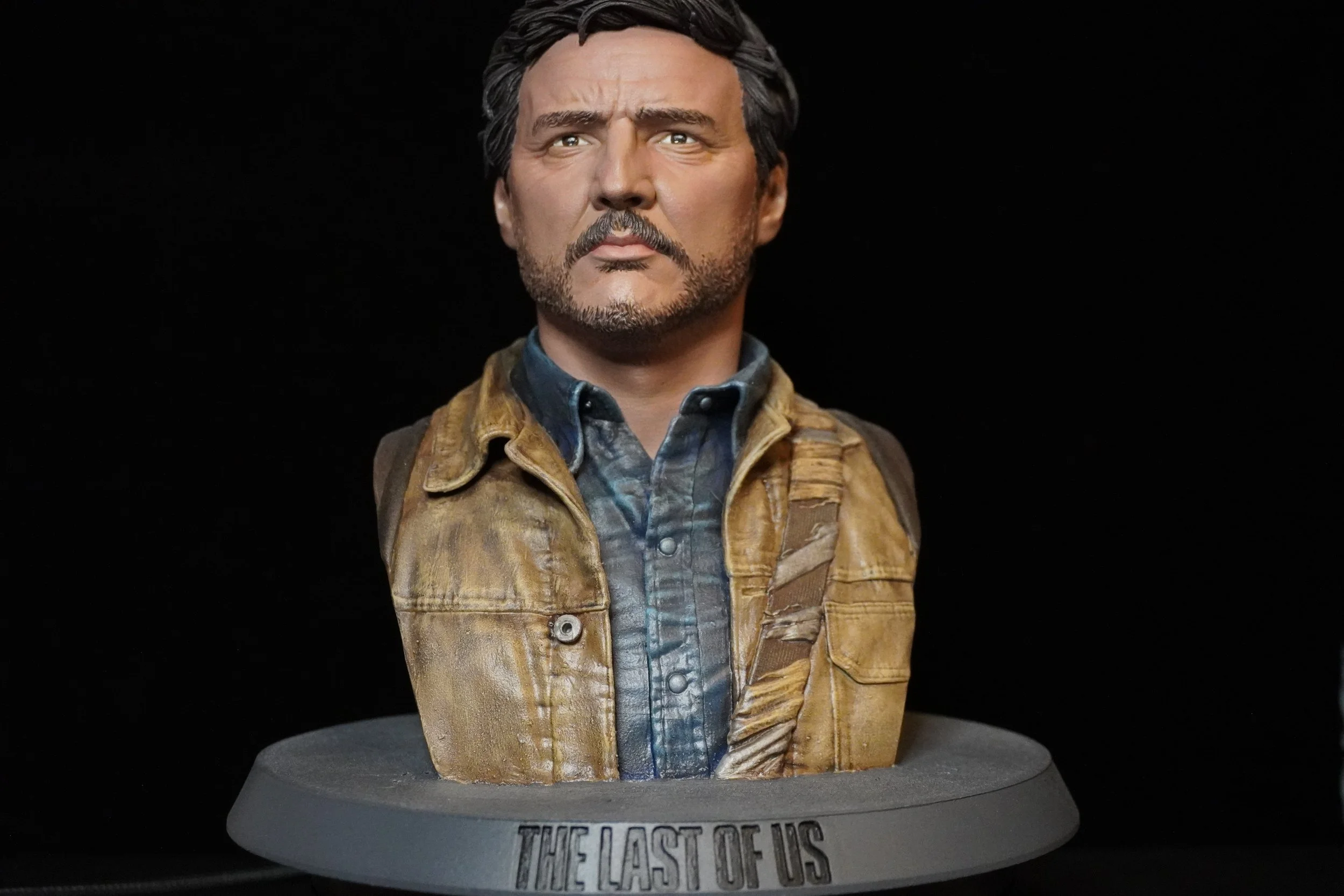 A detailed bust of a man with dark hair and a beard, wearing a denim shirt and a tan leather jacket, mounted on a stand labeled "The Last of Us" against a black background.