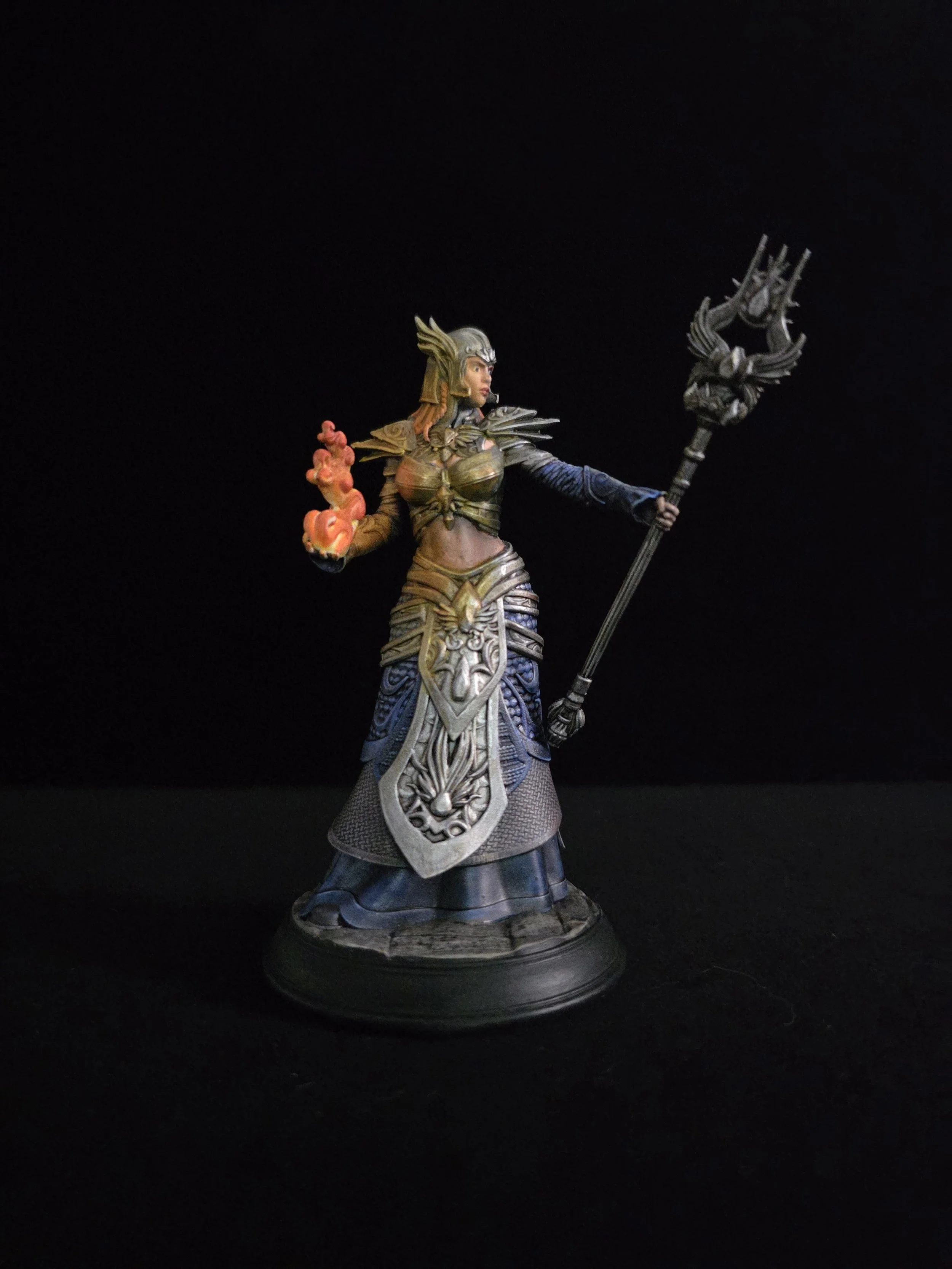 A detailed miniature fantasy figure of a female warrior with ornate armor, holding a staff in one hand and a flame in the other, standing on a round black base against a dark background.
