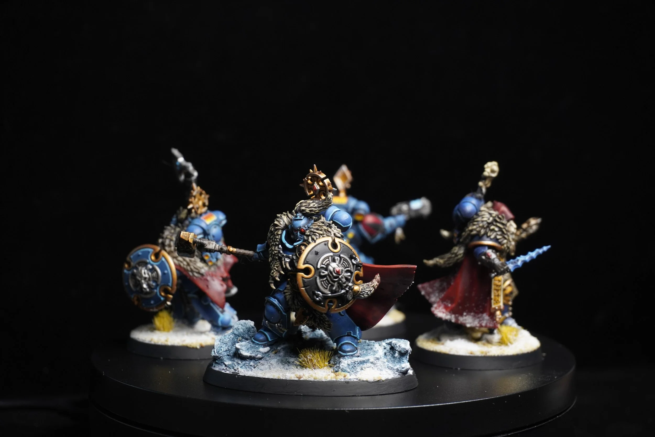 Four painted fantasy miniature warriors in armor, wielding weapons, standing on a black circular display base with snowy terrain, against a black background.