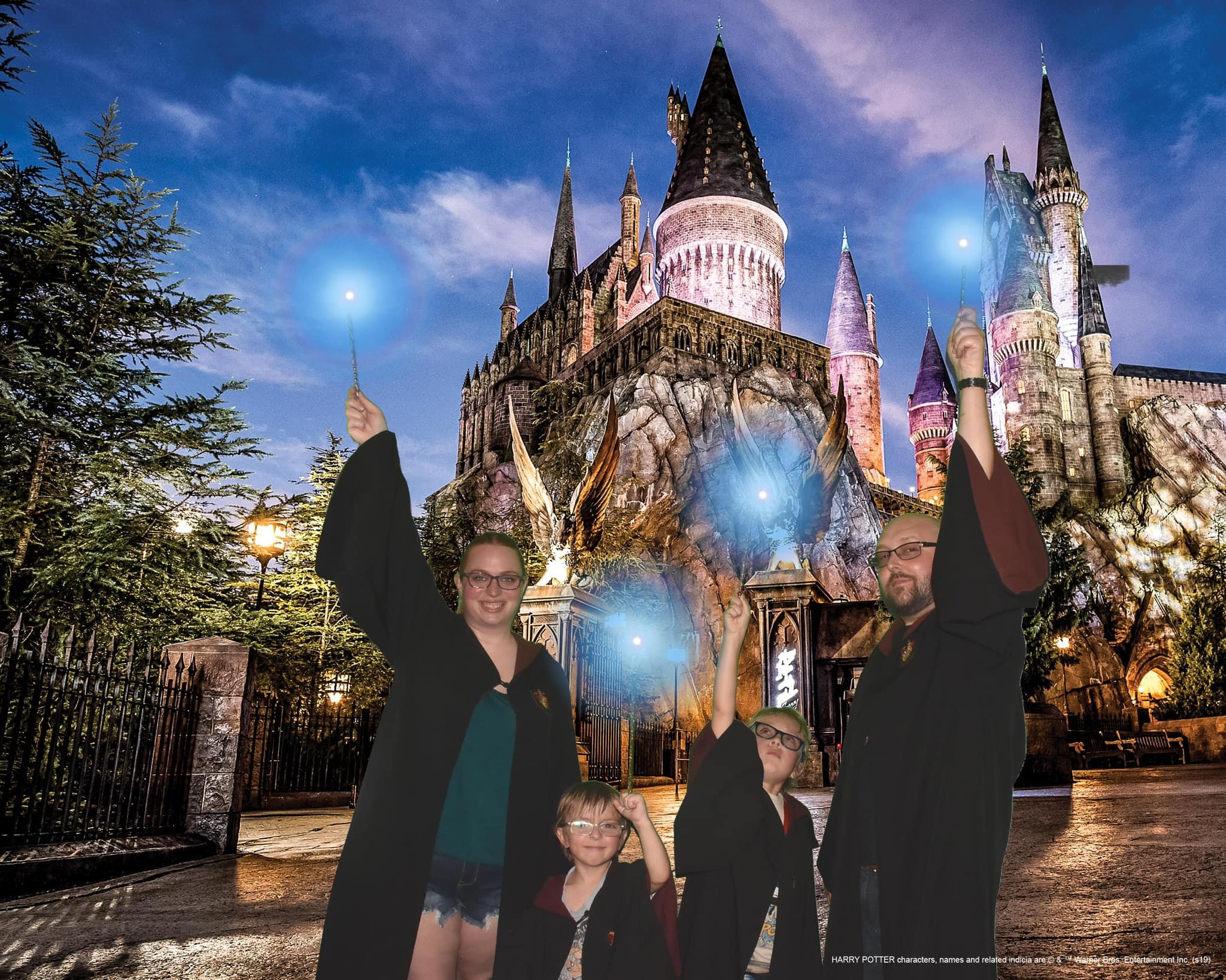 Family dressed in Hogwarts robes, standing in front of an illuminated Hogwarts castle with wands pointed into the air, nighttime sky in the background.
