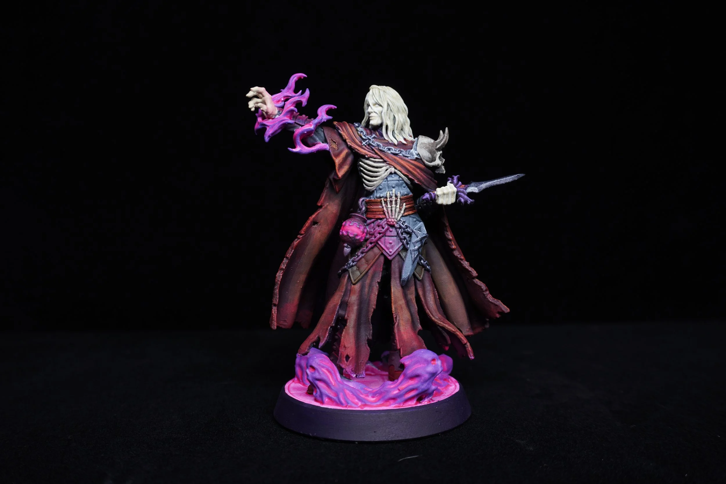 A detailed miniature figurine of a fantasy character with long blond hair, wearing a tattered red and grey cloak, holding a dagger, with purple flames emanating from the hand, standing on a base decorated with pinkish-purple swirling mist.