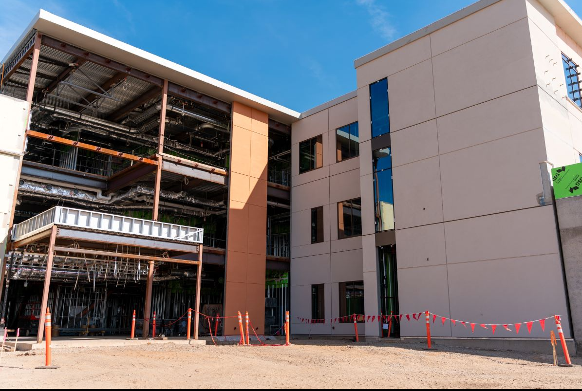 Construction site of a modern building with exposed framework and partially finished exterior walls, orange safety cones and fencing, and clear blue sky.