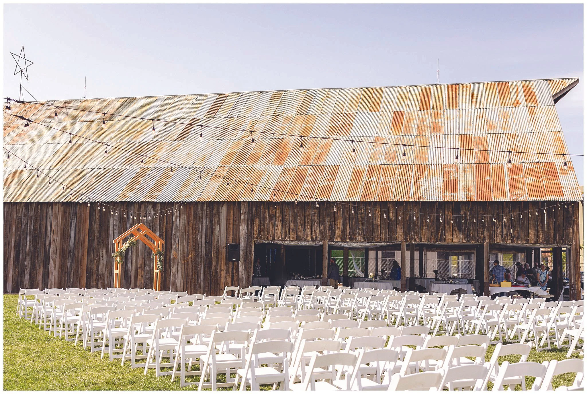 Outdoor wedding ceremony setup with rows of white chairs facing a rustic barn decorated with string lights and a wooden arch, on a grassy area.