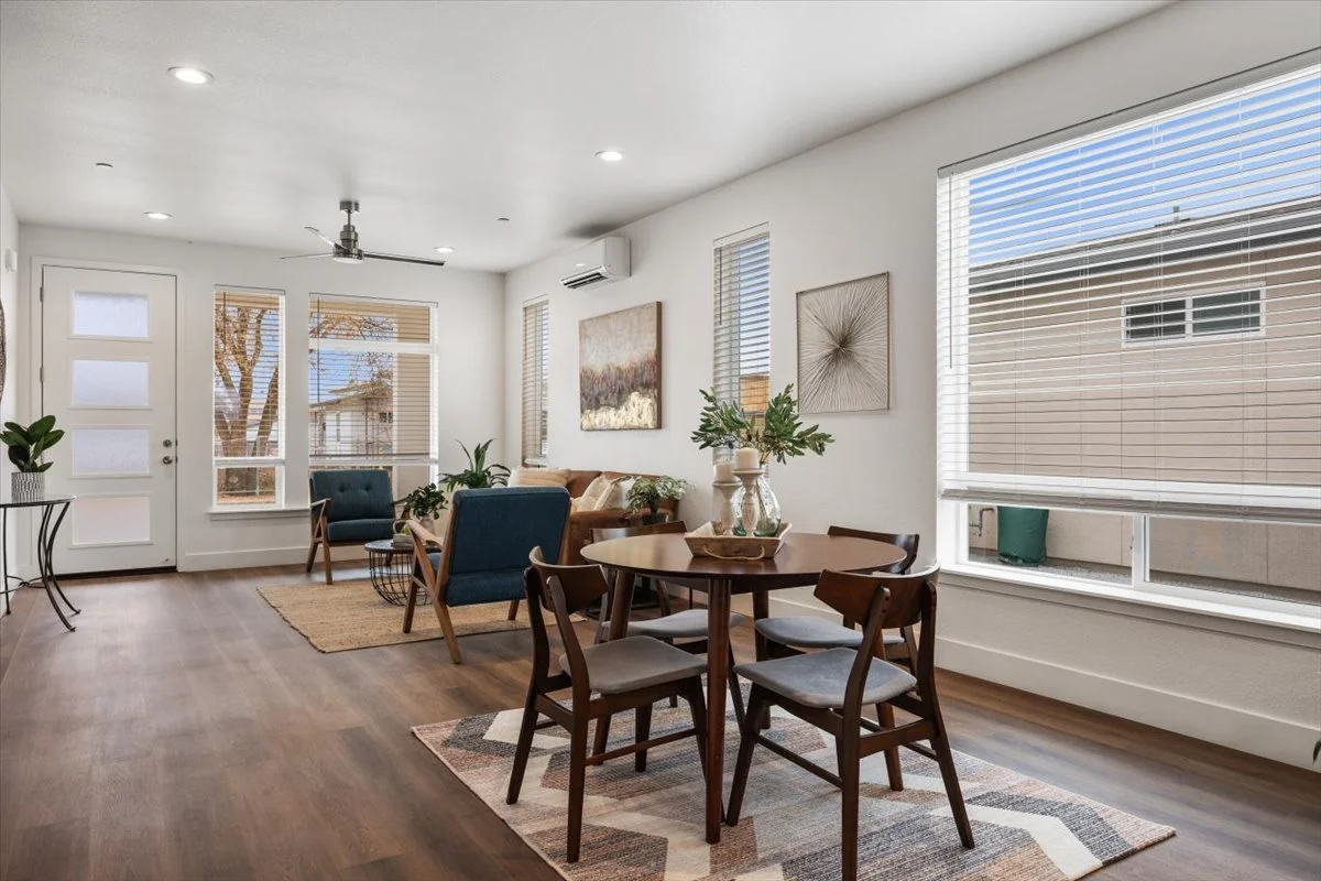 Living and dining room with large windows, hardwood floors, a round dining table with four chairs, a seating area with a sofa and chairs, indoor plants, wall art, and a door with glass panels.