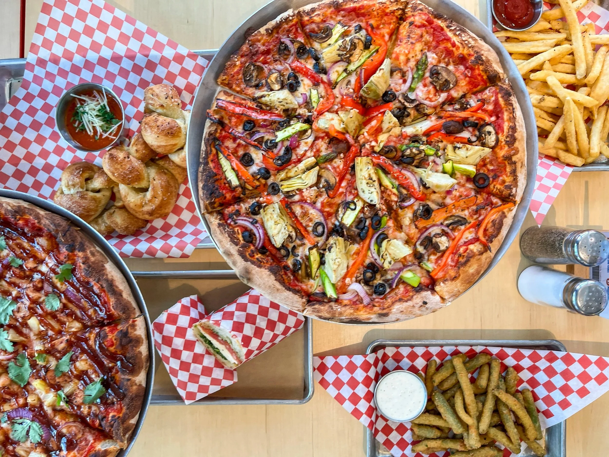 A table with two pizzas, one with vegetables and olives, another with chicken and cilantro, along with side dishes of garlic knots, French fries, and dipping sauces, topped with salt and pepper shakers.