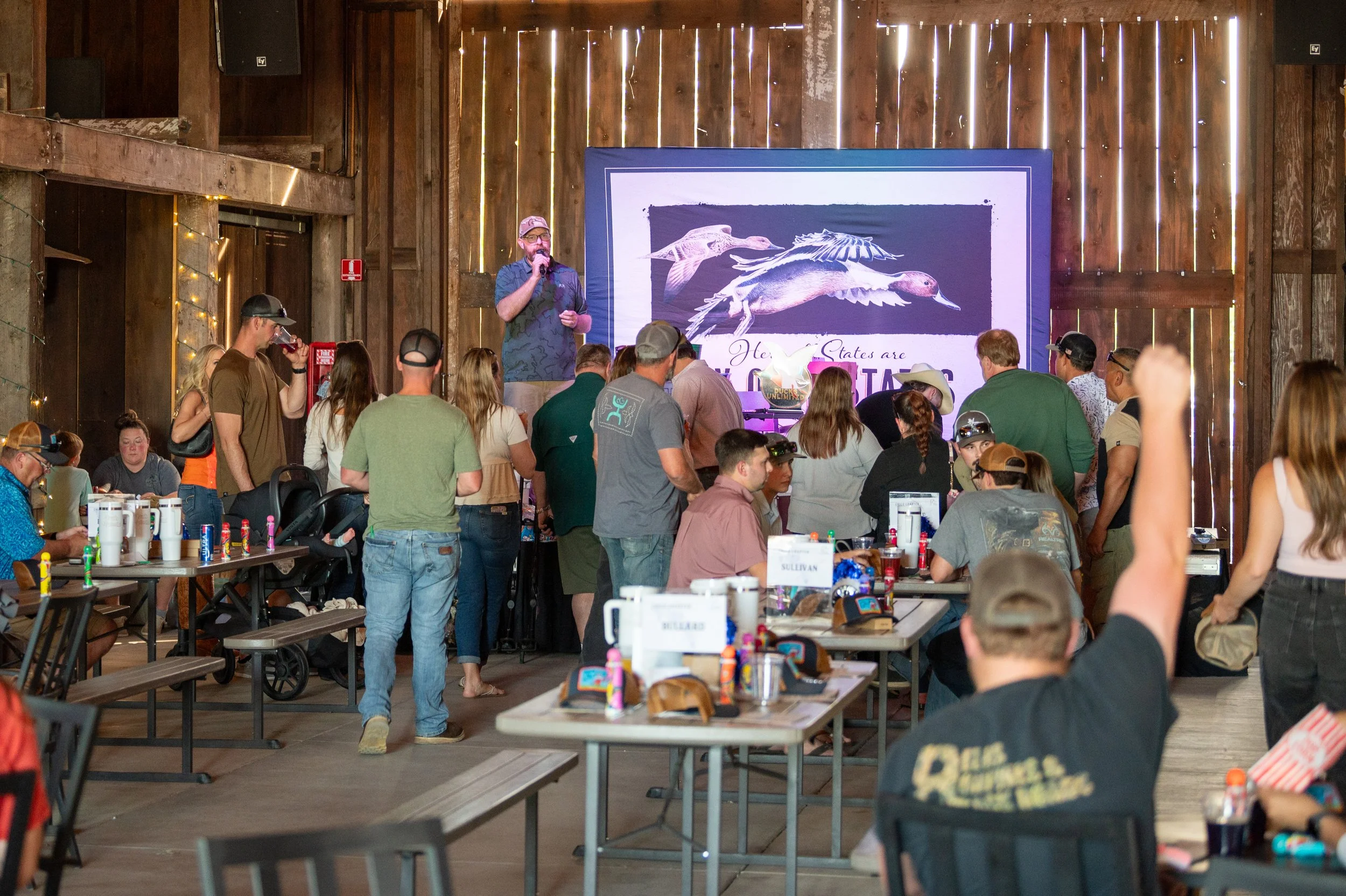 A gathering of people in a rustic barn-style venue. A man is on stage speaking into a microphone, in front of a large screen displaying birds and text. Attendees are standing, sitting, drinking, and engaging in conversation, with some wearing hats an