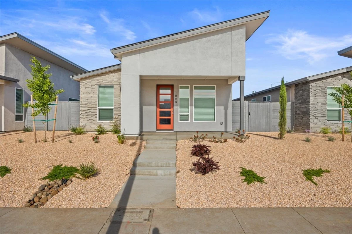 Front view of a modern house with a minimalist landscape, including small trees, shrubs, and rocks, and an orange front door.