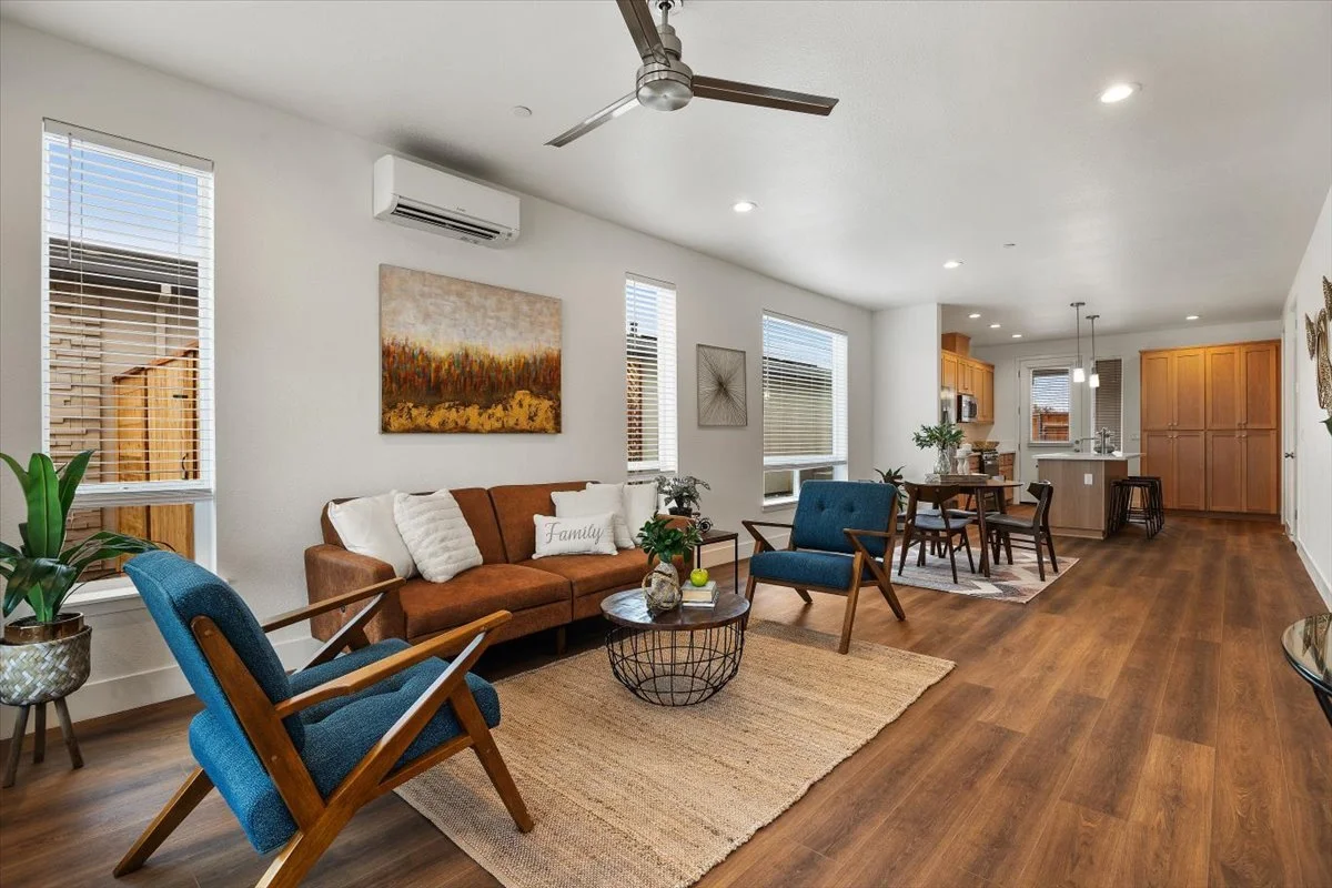 Open-concept living room and kitchen with hardwood floors, white walls, and modern furniture, including a brown sofa, blue armchairs, and a dining table.