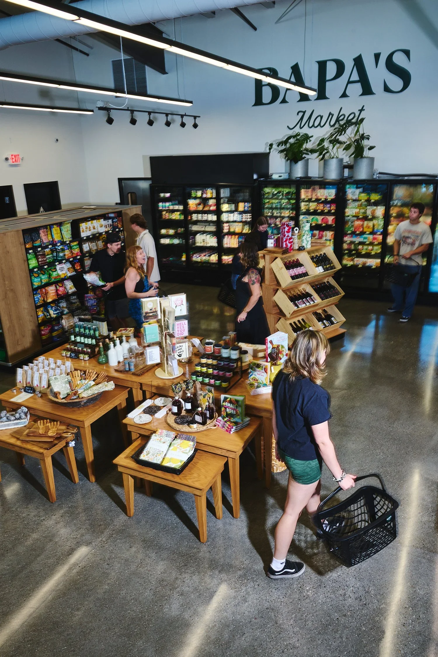 Interior of a grocery store named BAPA'S Market, with customers browsing shelves of snacks, beverages, and assorted products on display tables and refrigerated sections.
