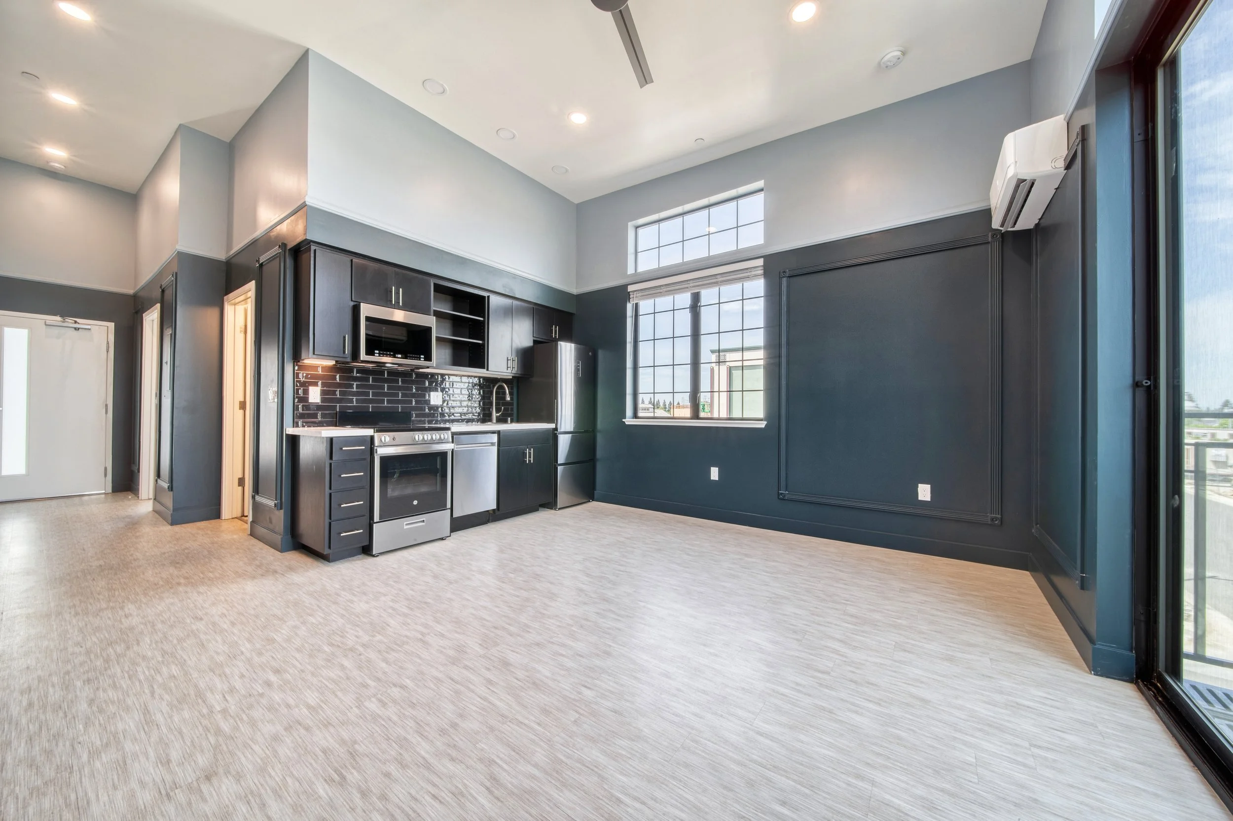 Empty modern kitchen with dark cabinets, stainless steel appliances, black subway tile backsplash, large window, high ceiling, and light wood flooring.