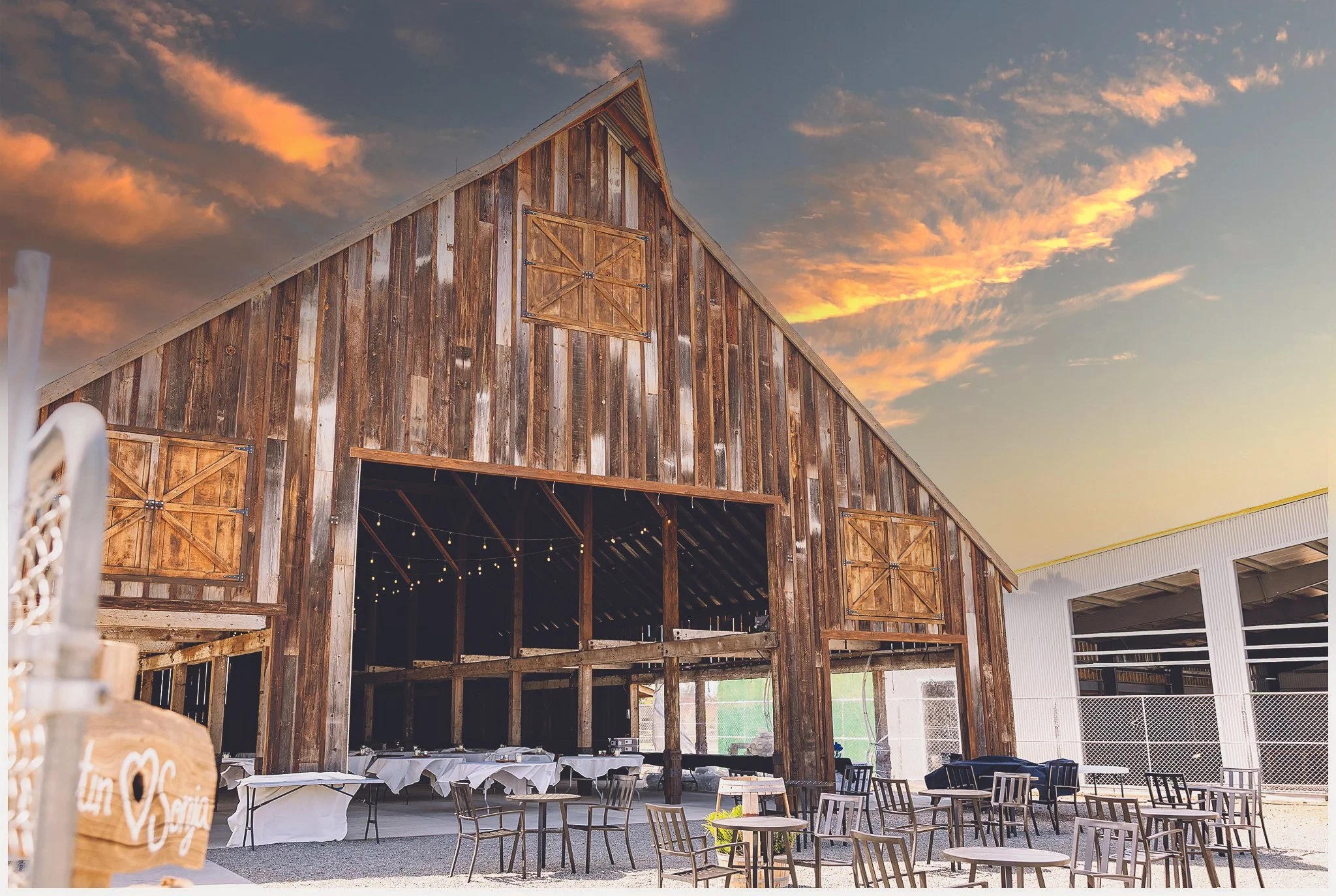 A rustic outdoor setting featuring a large wooden barn with a sloped roof and barn doors, decorated with string lights, at sunset with a partly cloudy sky.