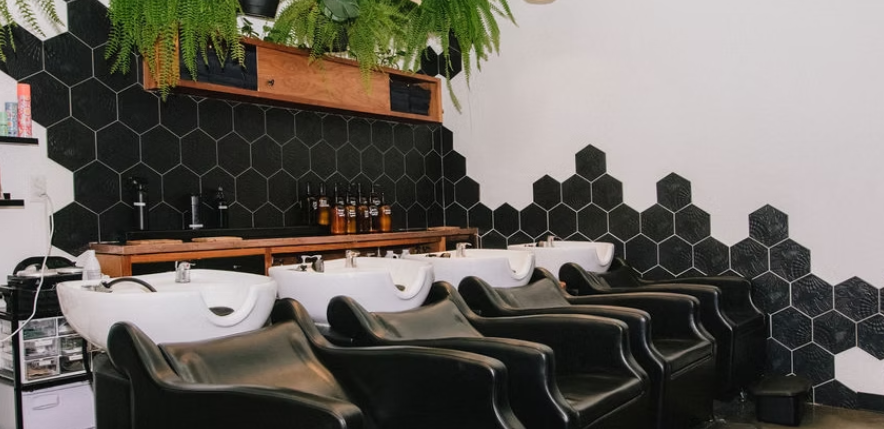 Salon with black massage chairs in front of hair washing sinks, black hexagon tile wall, with green hanging plants and a wooden shelf with bottles.